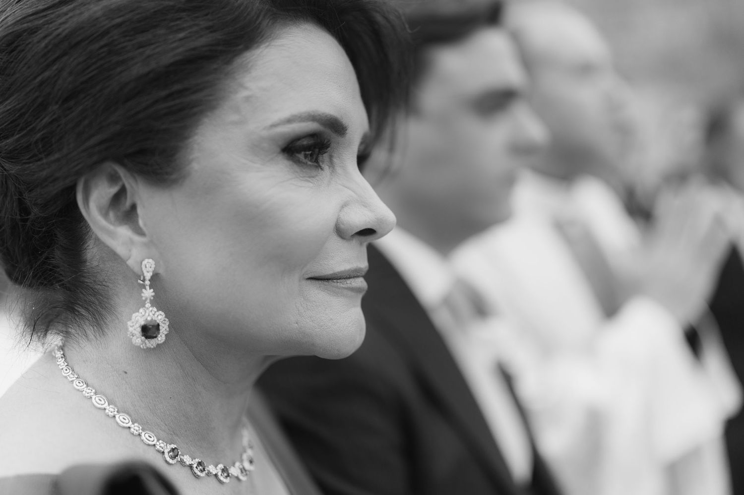 Black and white close-up of elegant woman wearing diamond necklace and earrings at wedding.