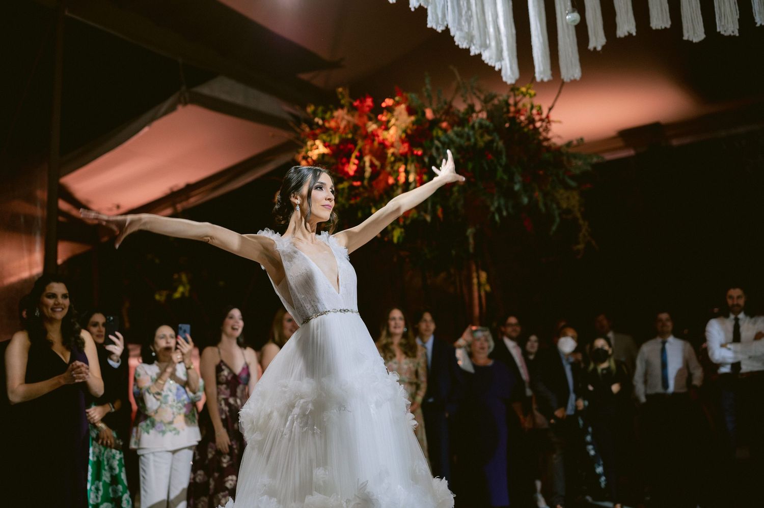 Bride in white tiered gown dances with arms raised before vibrant floral arrangement and applauding guests.