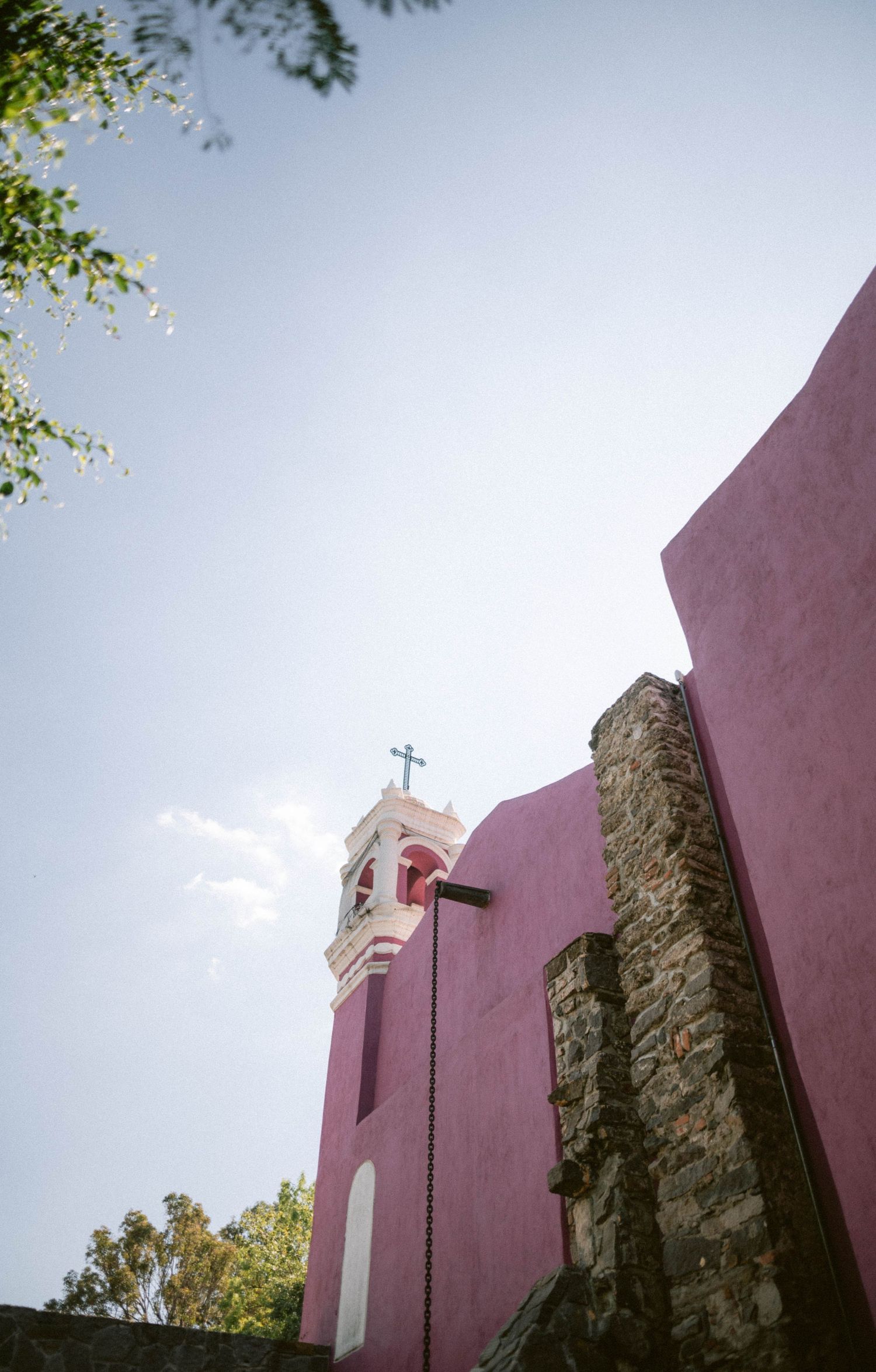 Low angle view of pink colonial church with white bell tower and cross against blue sky.
