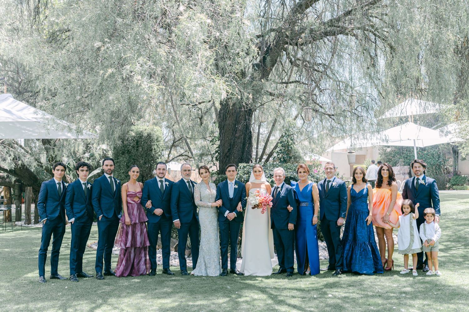 Large wedding party poses together under sweeping willow tree in elegant outdoor garden setting.