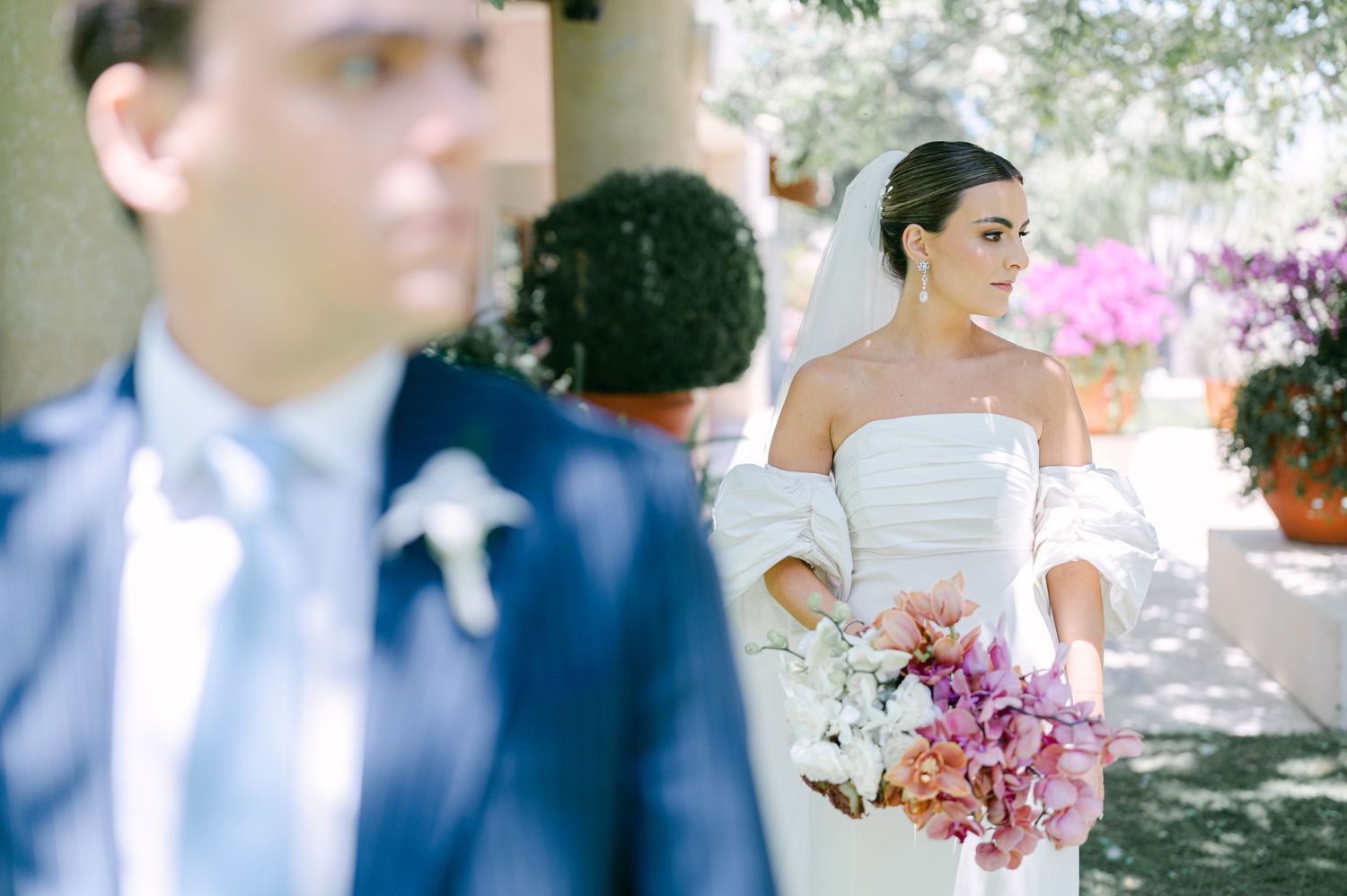 Bride in off-shoulder wedding gown holding pink floral bouquet stands behind groom in navy suit at outdoor venue.