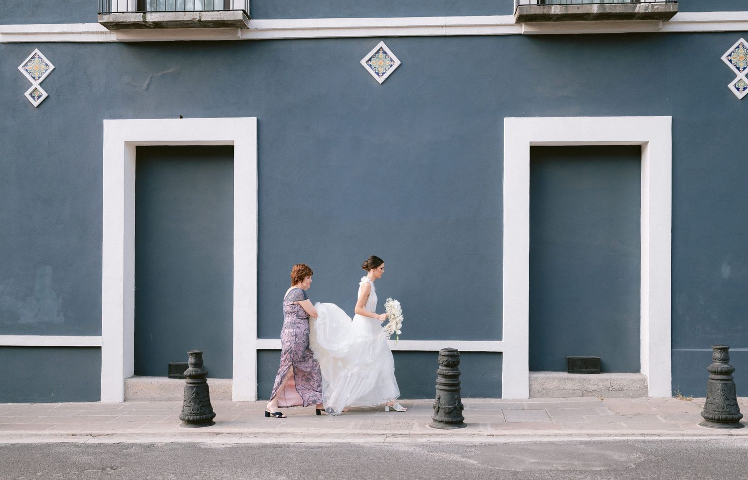 Bride in white gown walking along a blue building exterior with attendant carrying her train.