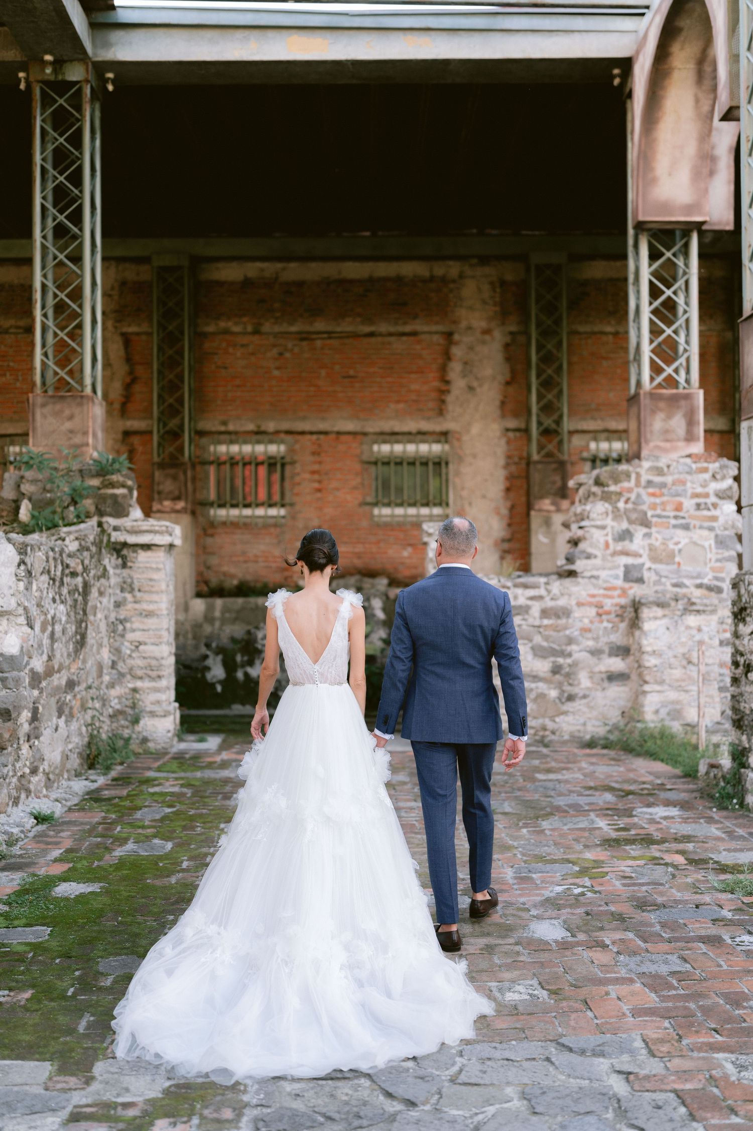Bride in backless tiered tulle gown and groom in navy suit walking through historic brick ruins.