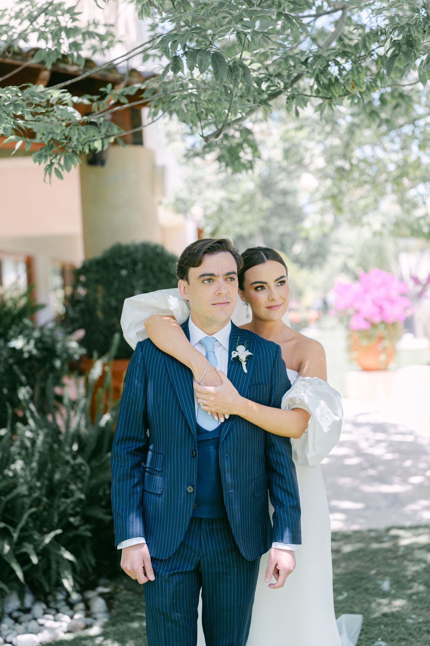 Bride in white off-shoulder gown embraces groom in navy pinstripe suit outdoors with pink flowers in background.