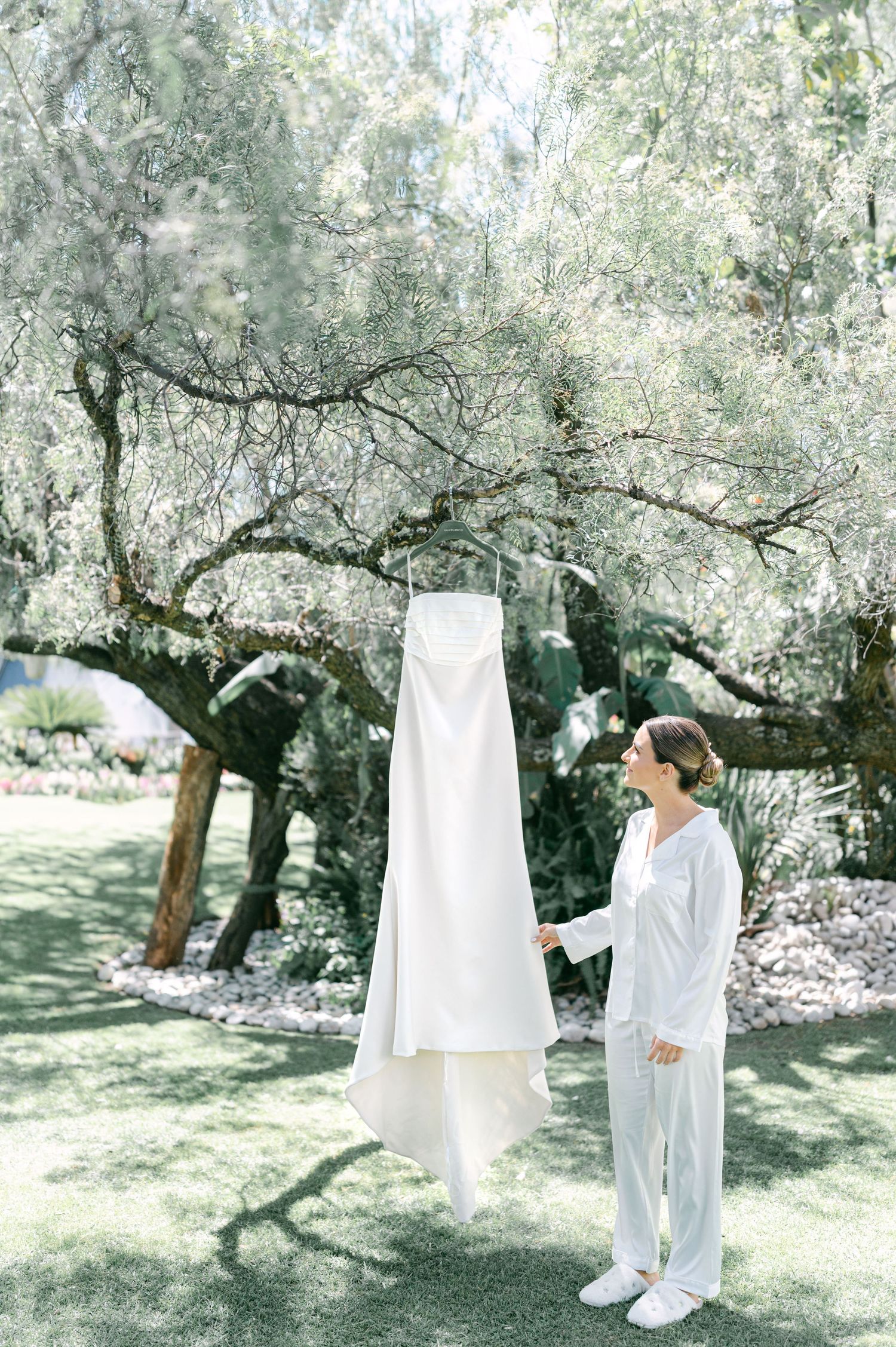 Bride in white pajamas admiring her strapless wedding gown hanging from an olive tree in garden.
