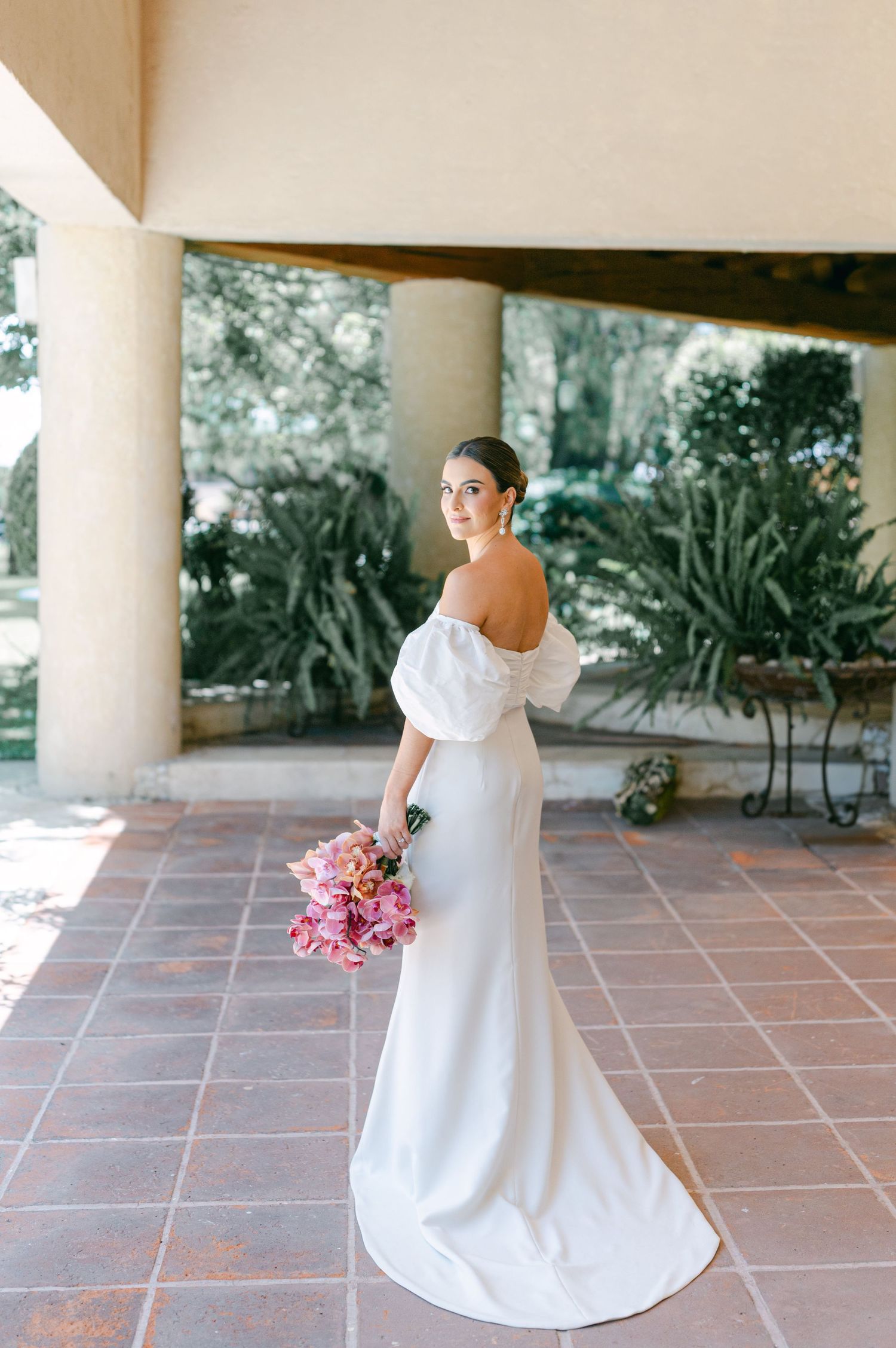 Bride in sleek white off-shoulder gown with train looks back over shoulder while holding pink floral bouquet.