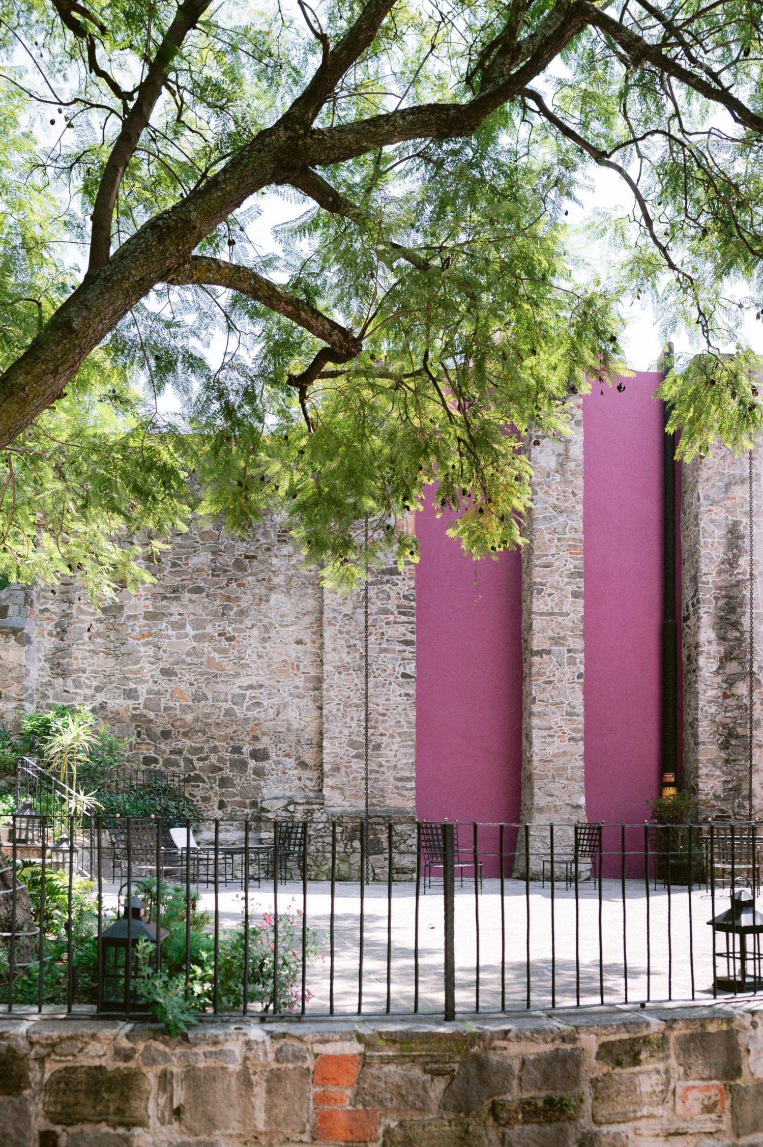 Pink painted columns against a rustic stone wall with iron fence and green trees in a Mexican courtyard.