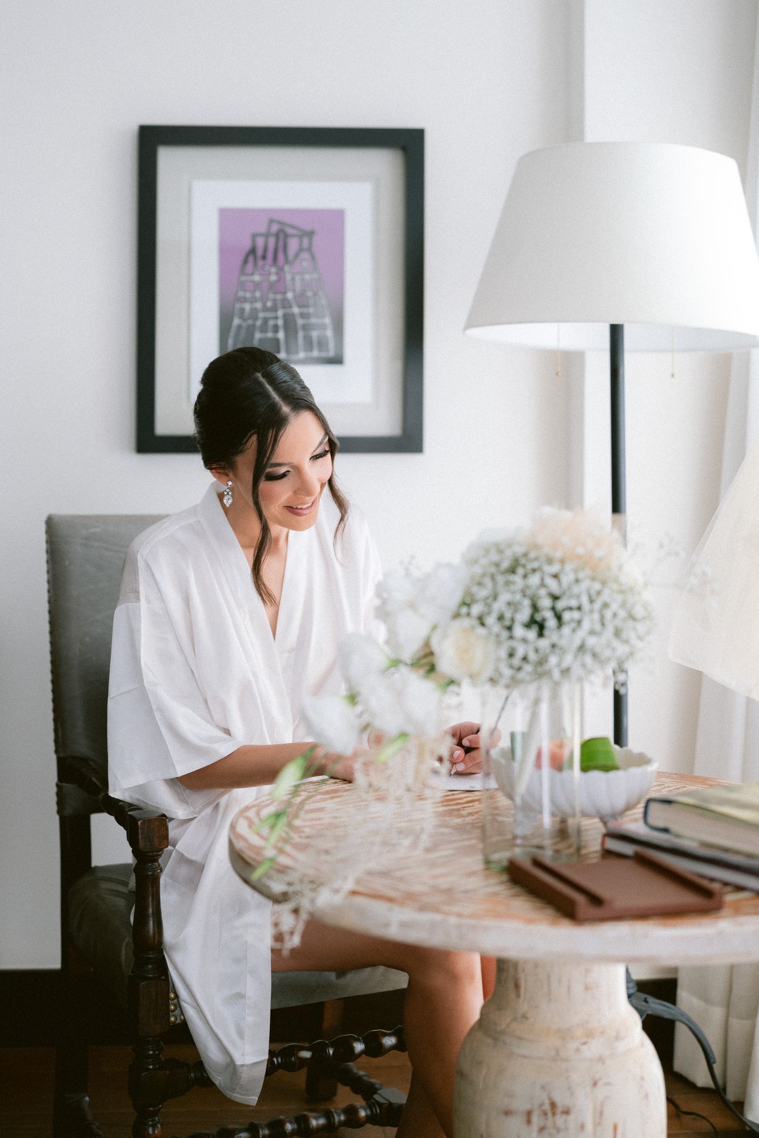 Smiling bride in white robe sitting at table with white floral arrangements on wedding morning.