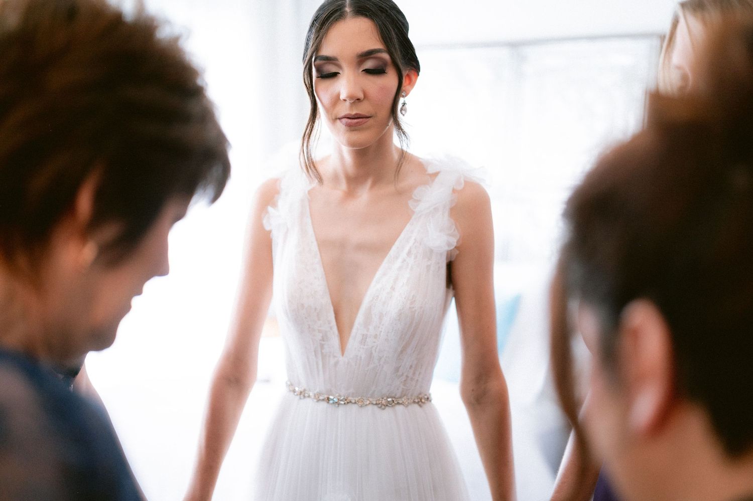 Bride in elegant deep V-neck white wedding gown with feather straps being assisted by attendants.