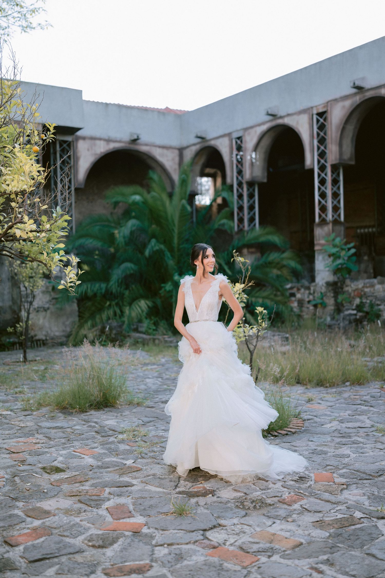 Bride in layered tulle ball gown twirls in cobblestone courtyard of historic arched building with palm trees.