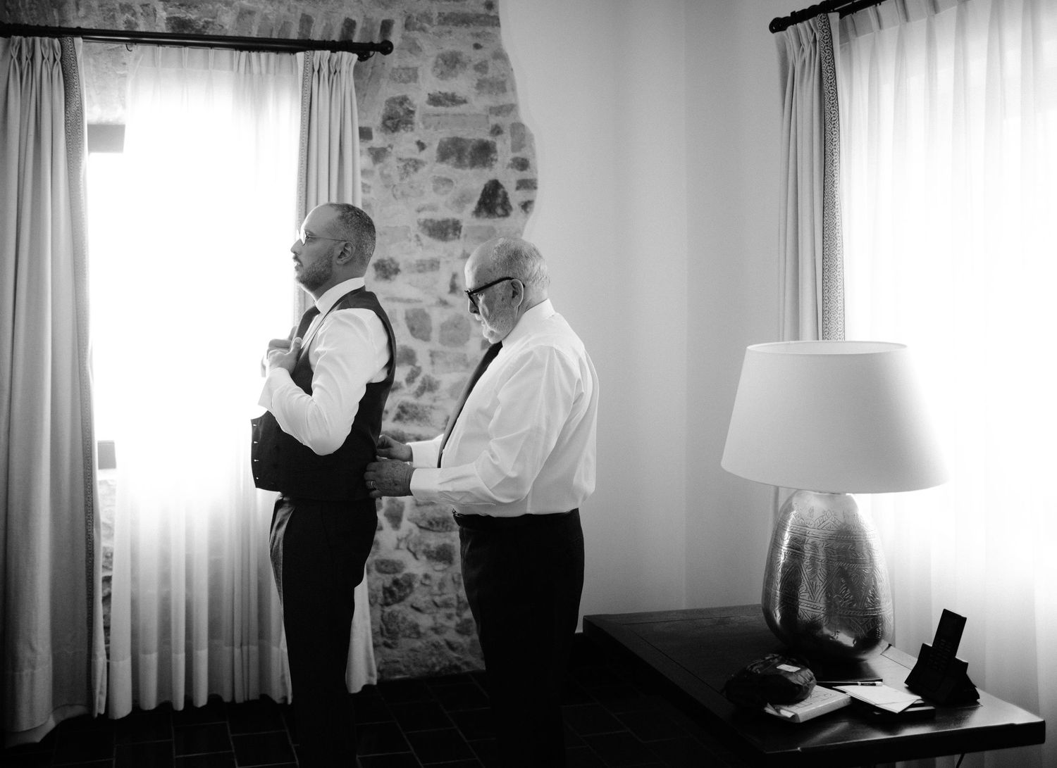 Black and white photo of groom being helped with tie by older gentleman in elegant room.