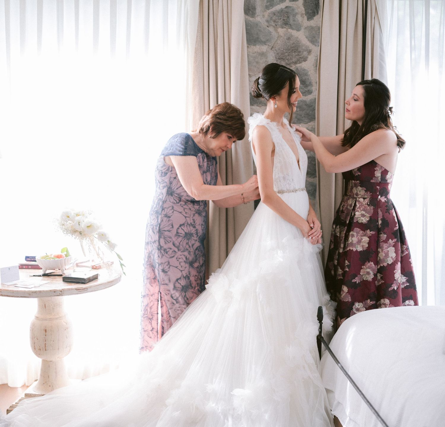 Two women help bride into her white wedding dress in a sunlit room.