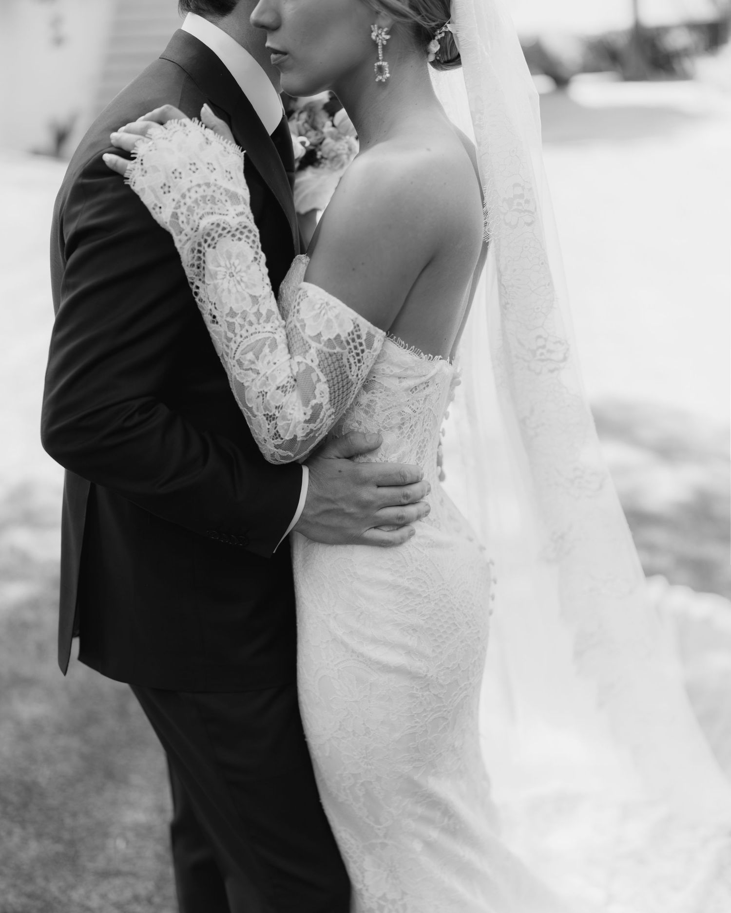 Black and white photo of bride and groom embracing, bride wearing lace off-shoulder gown with veil.