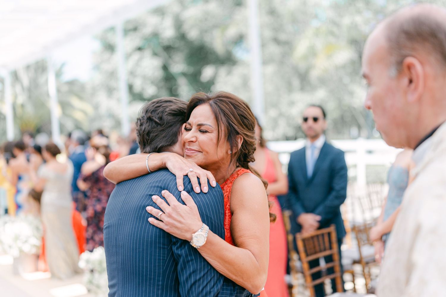 Woman in orange dress hugging a man in a blue suit at an outdoor wedding ceremony.