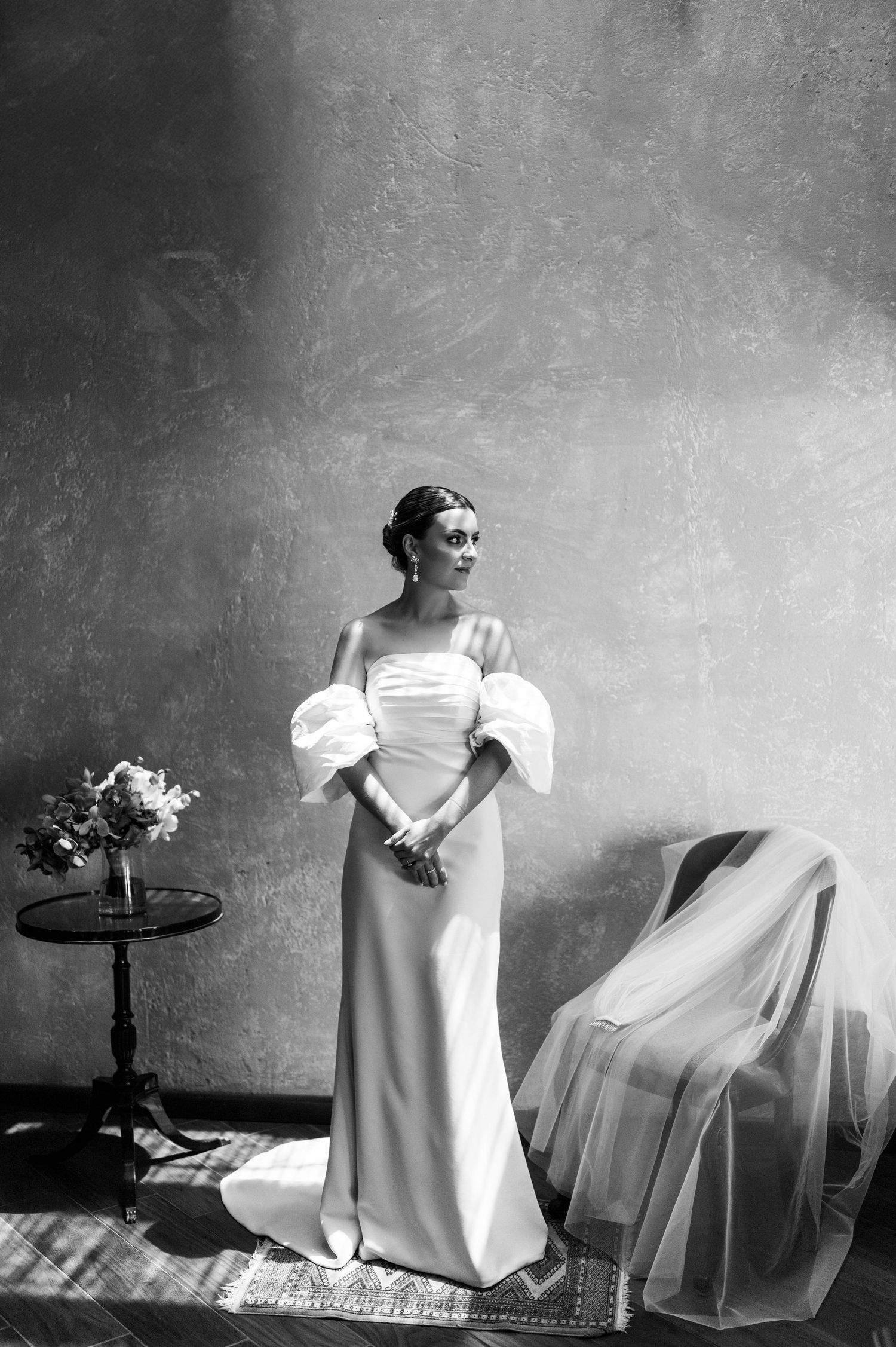 Elegant bride in off-shoulder white gown with puff sleeves stands in black and white portrait beside floral bouquet.
