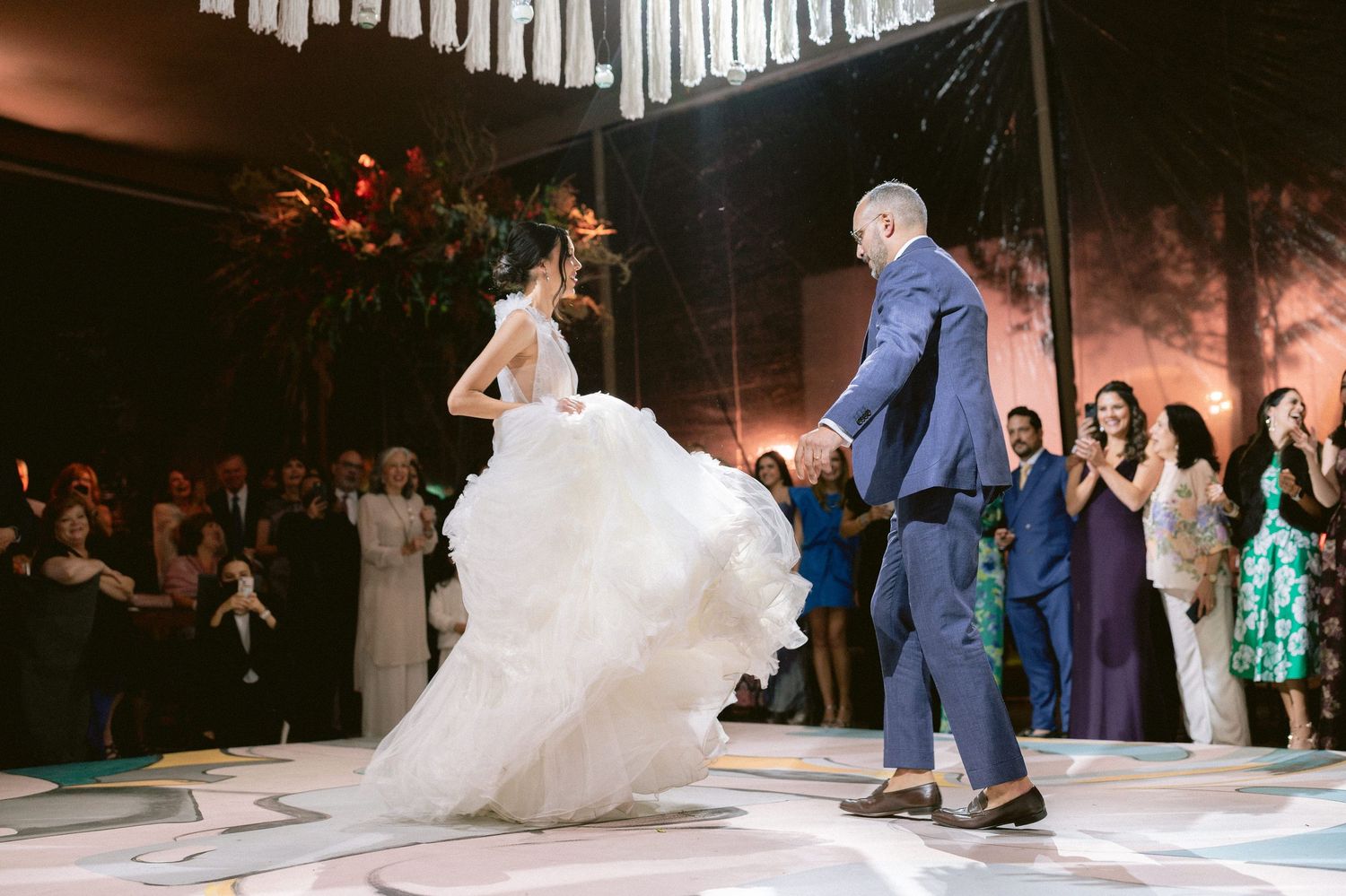 Groom spins bride in a voluminous white gown during their first dance at a lively wedding reception.