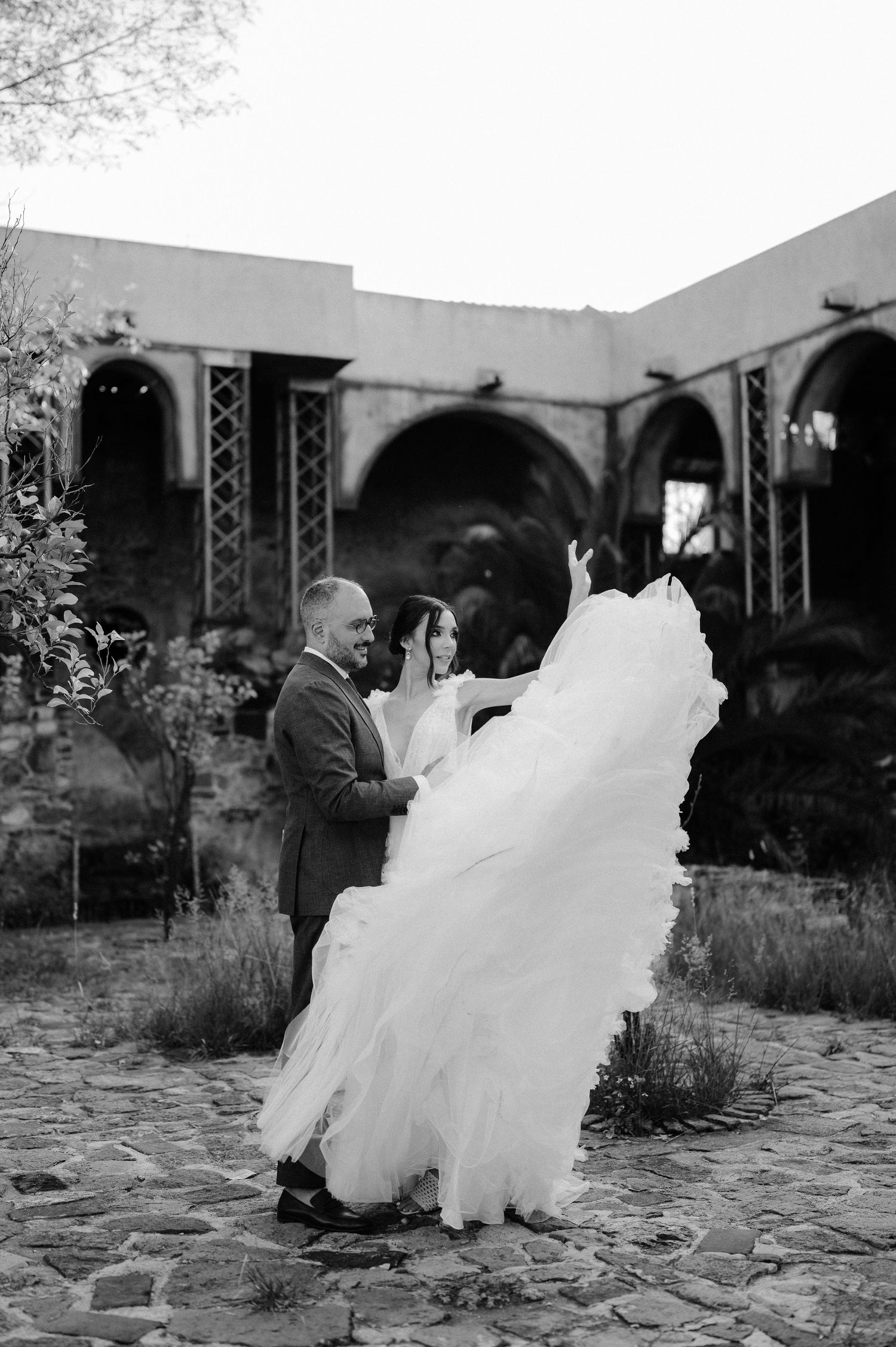 Black and white photo of couple dancing as brides dramatic tulle gown billows in historic stone courtyard.