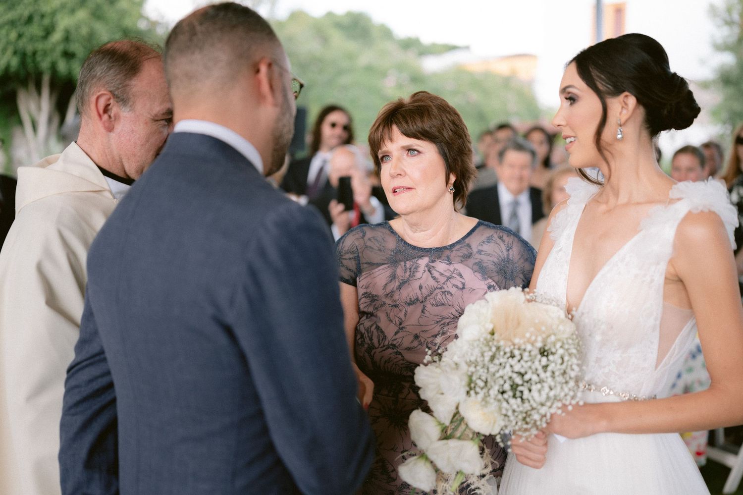 Bride holding white bouquet smiles as officiant speaks during outdoor wedding ceremony with guests.