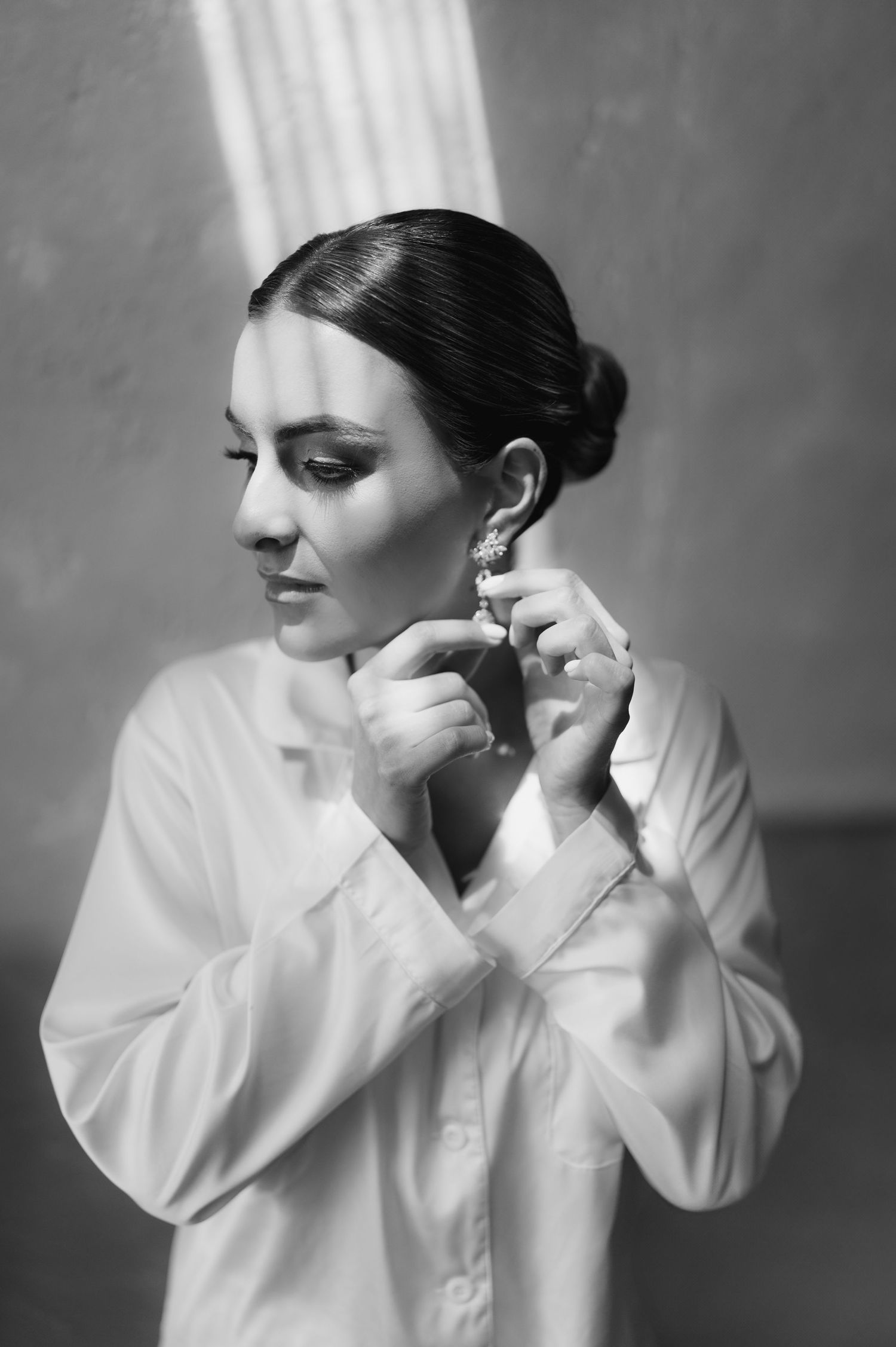 Elegant woman in white shirt putting on earrings in a black and white portrait photo.