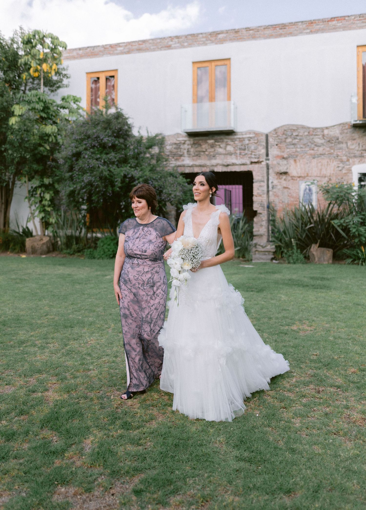 Bride in ruffled white gown walking with older woman across green lawn at rustic venue.