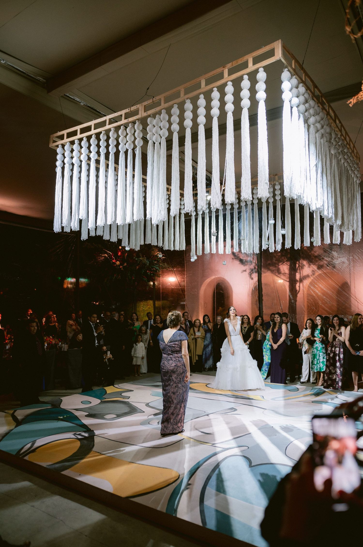 Bride and guest dance beneath stunning white macrame chandelier installation on artistic patterned dance floor.