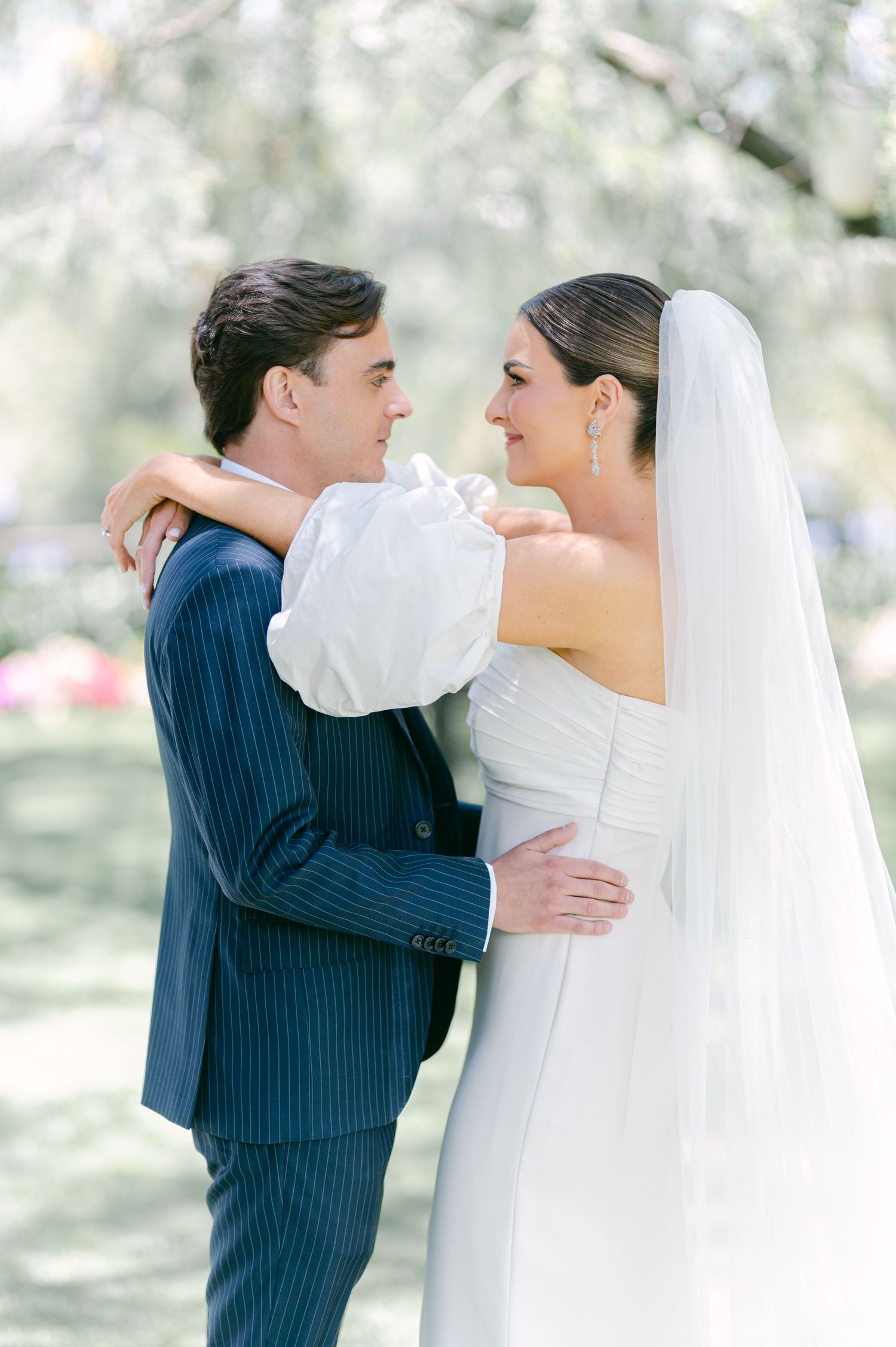 Bride and groom gazing at each other lovingly, she in white off-shoulder gown with veil, he in navy pinstripe suit.