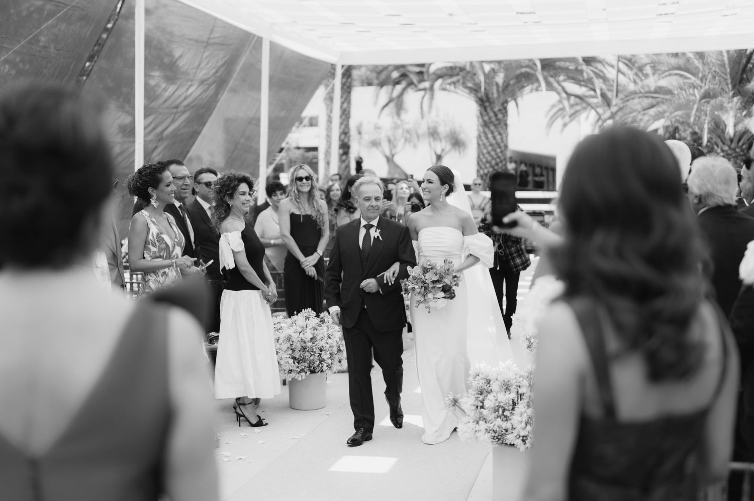 Black and white photo of bride walking down aisle with escort at outdoor wedding.