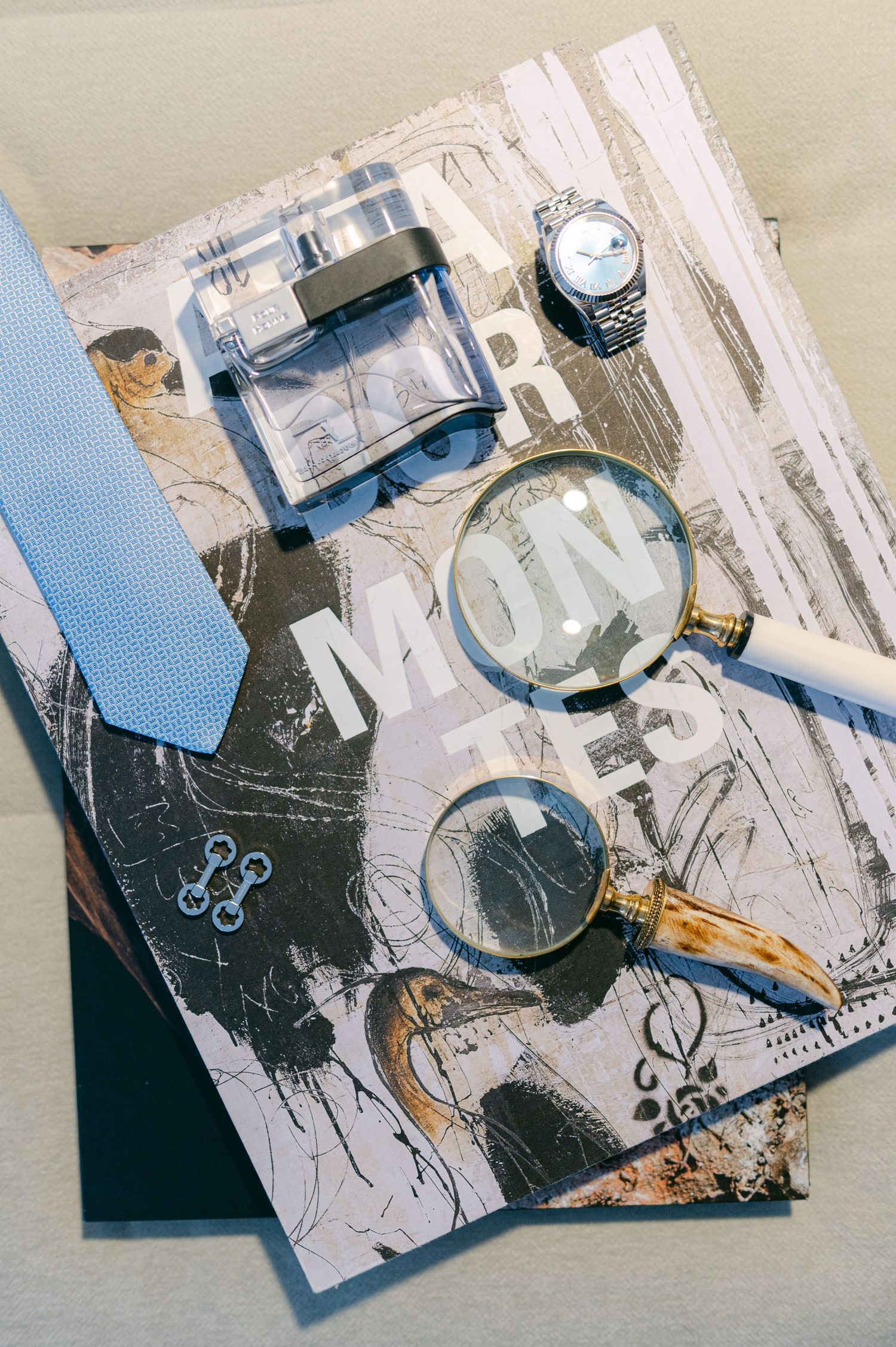 Groom's wedding flat lay with blue tie, cologne, watch, cufflinks, and magnifying glass on decorative paper.