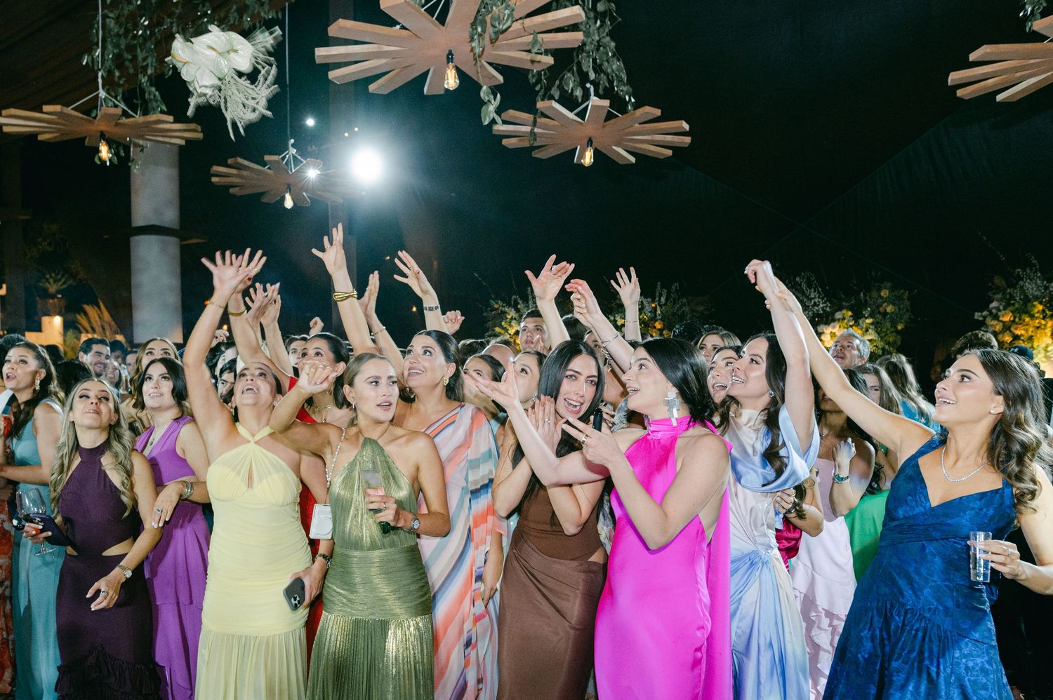 Colorful wedding guests in vibrant gowns reach up to catch the bridal bouquet toss at an outdoor evening reception.