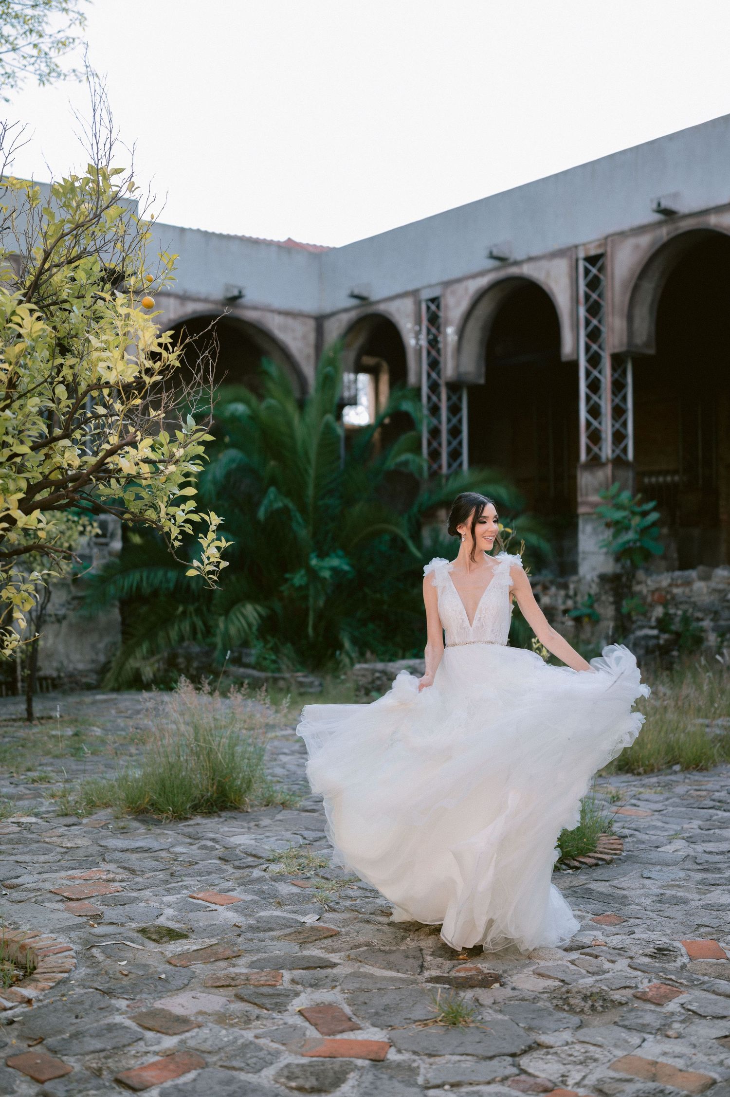 Bride in flowing white tulle wedding gown spins gracefully on cobblestone courtyard of rustic arched estate.
