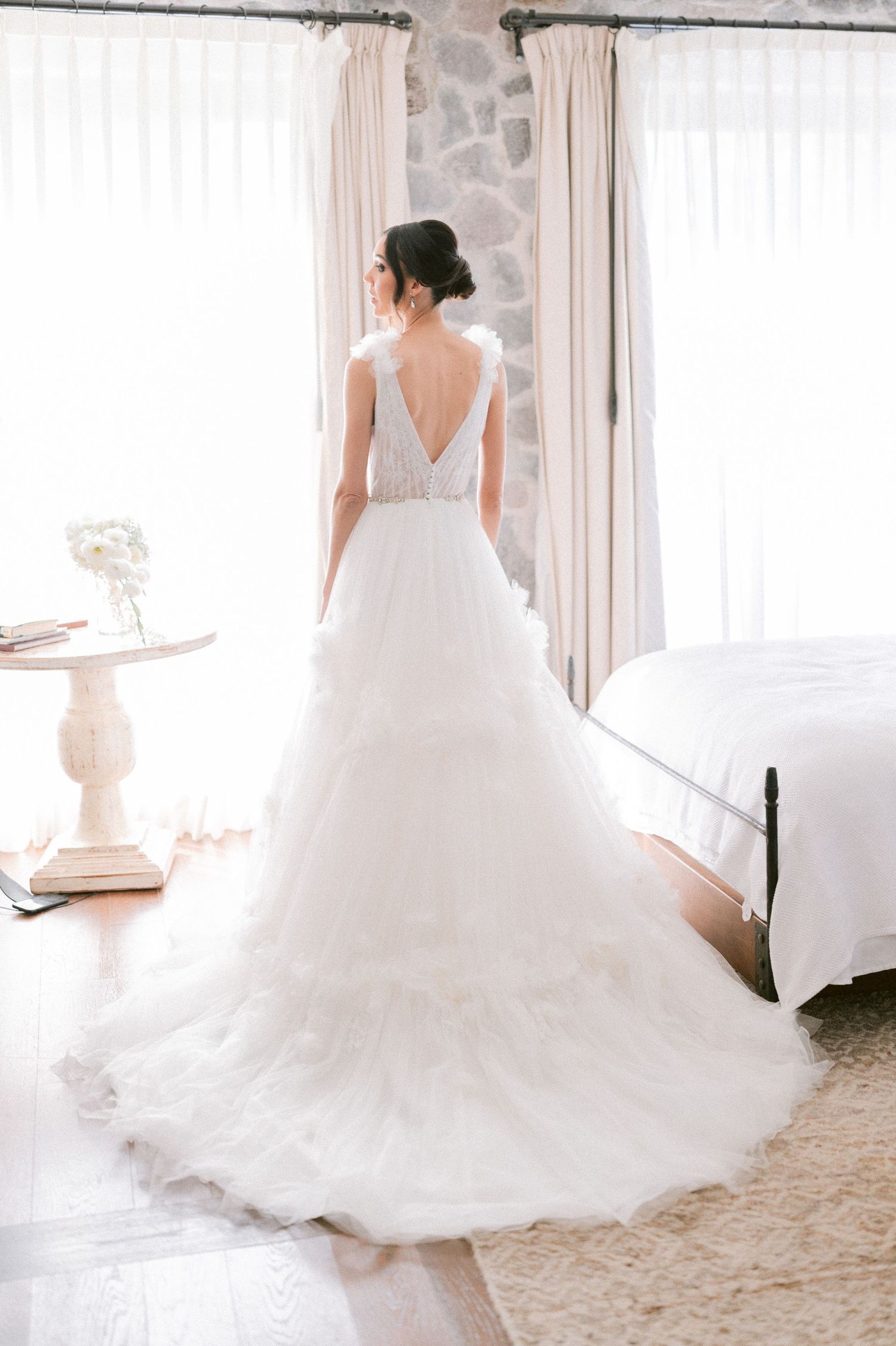 Bride in backless layered tulle wedding gown with cathedral train stands by window.