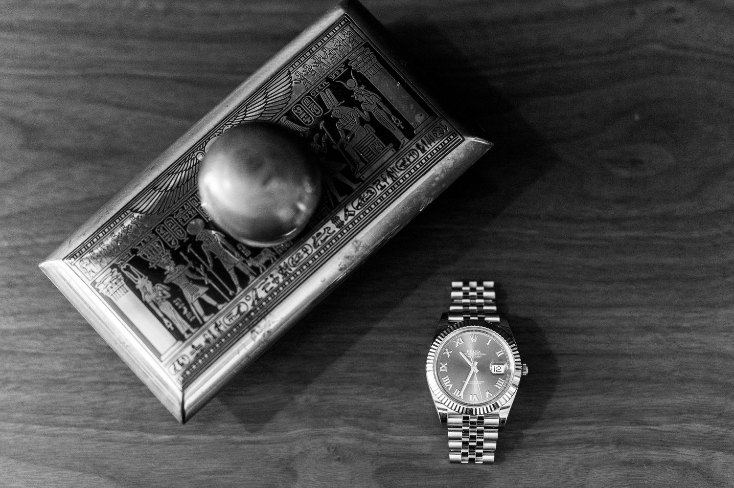 Black and white flat lay of a Rolex watch beside an ornate decorative jewelry box on wood surface.