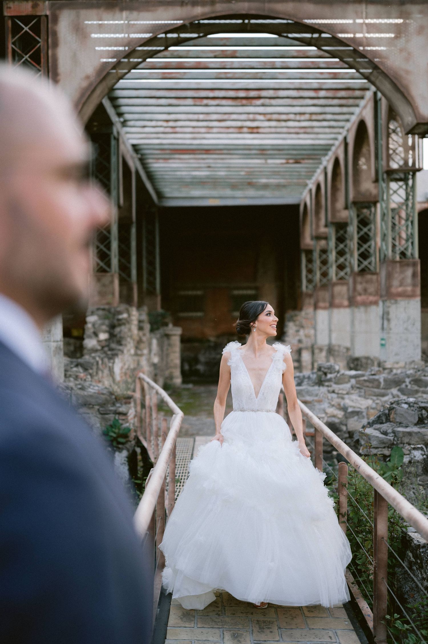 Bride in layered tulle gown stands on rustic bridge in blurred foreground of groom at historic venue.