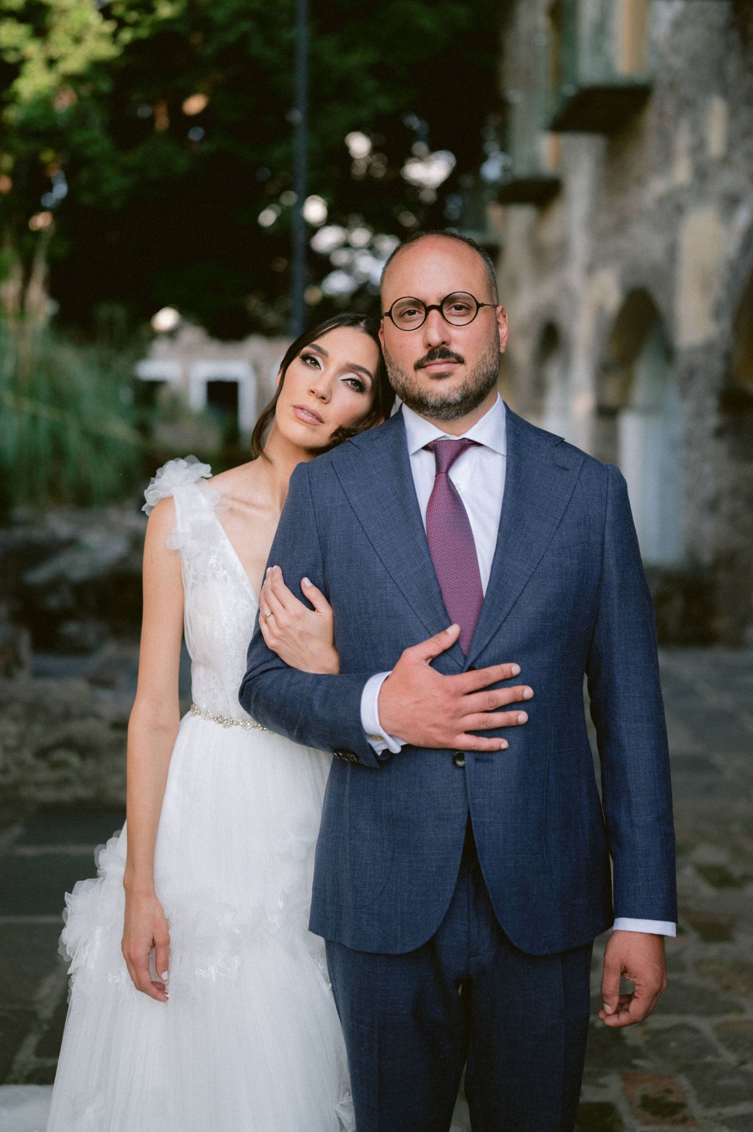 Bride in white tulle gown embraces groom in blue suit with purple tie at outdoor wedding venue.