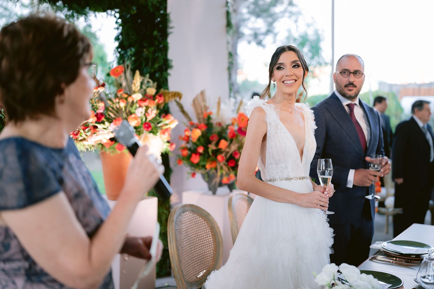 Smiling bride in a deep V-neck wedding gown listens to a toast while holding a champagne glass at reception.