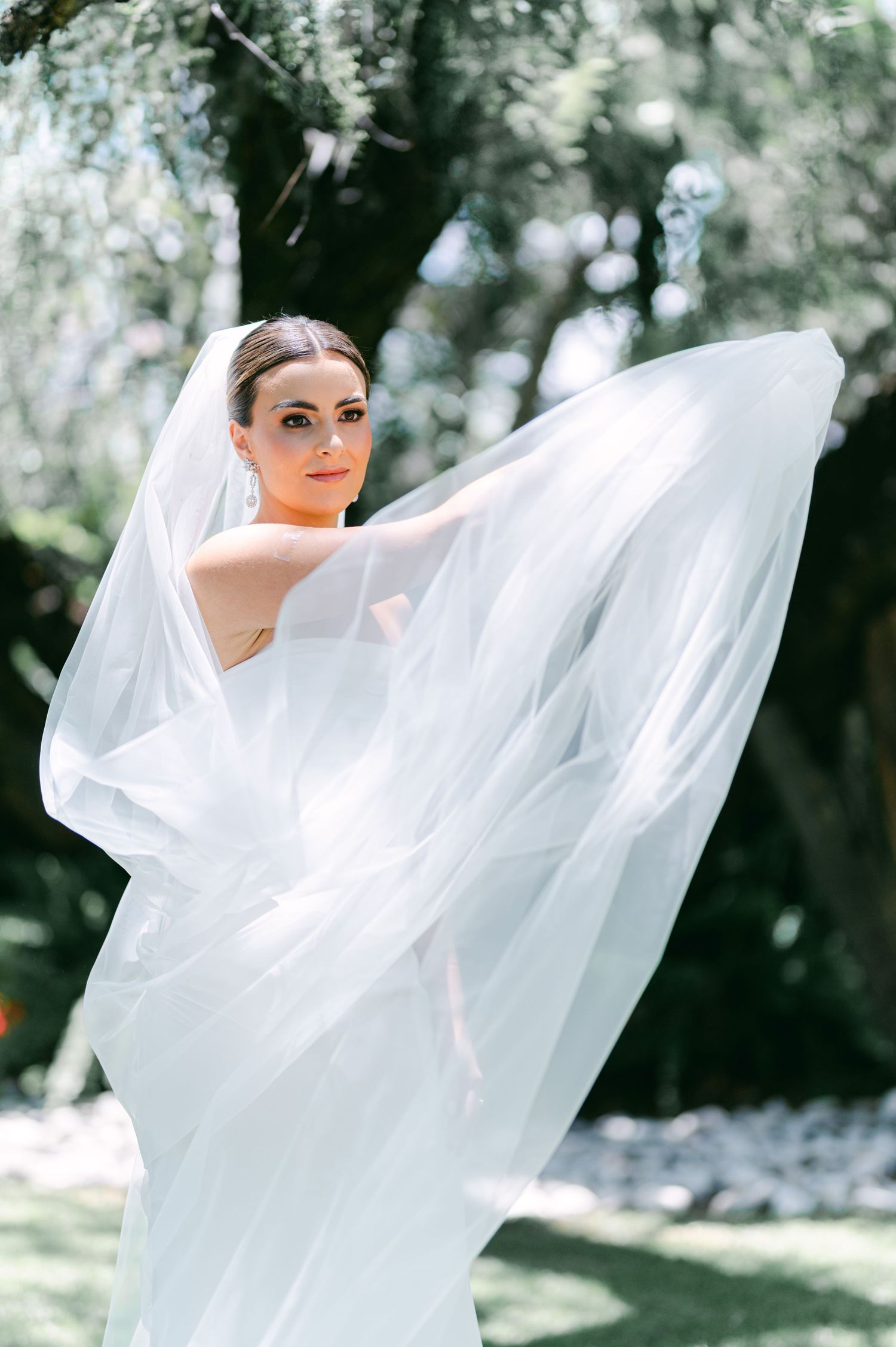 Beautiful bride twirls flowing veil in sunlit garden wearing sleek white wedding dress.
