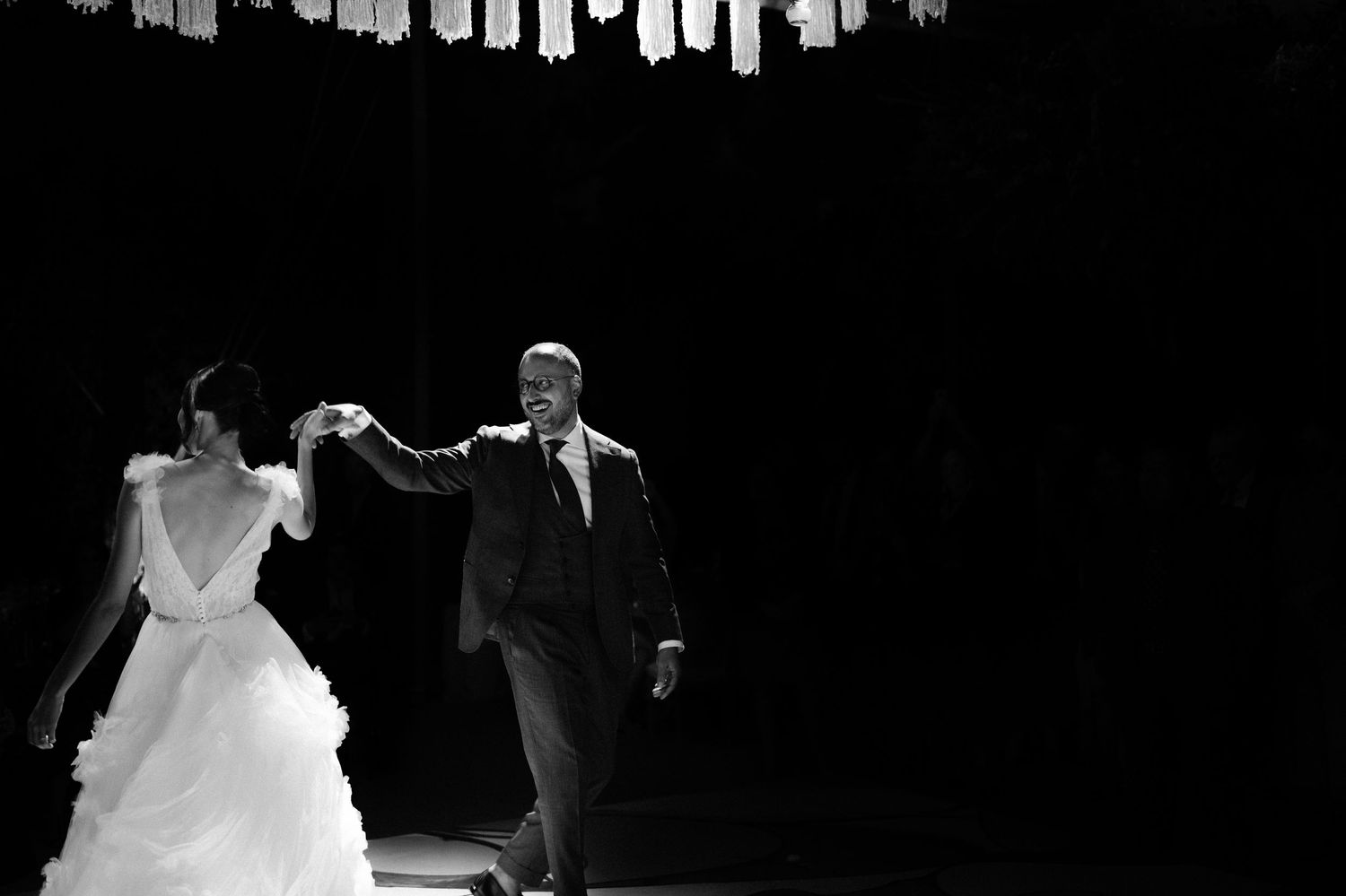 Black and white dramatic photo of bride and groom walking hand in hand through a spotlight at their reception.