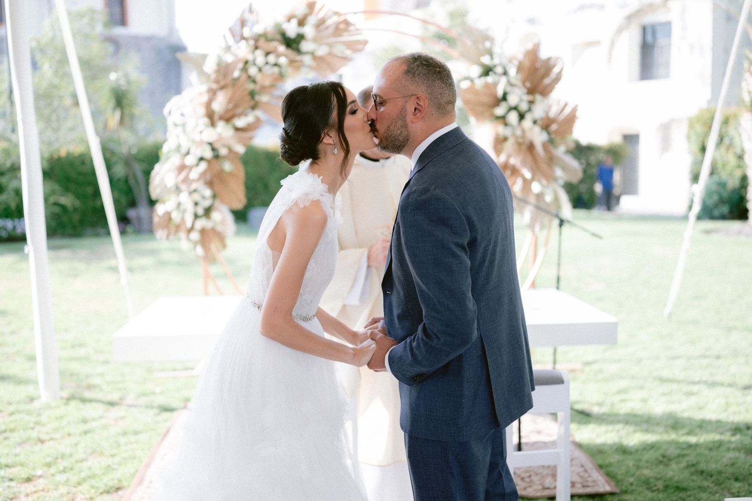 Bride and groom share first kiss under floral arch at beautiful outdoor garden wedding ceremony.