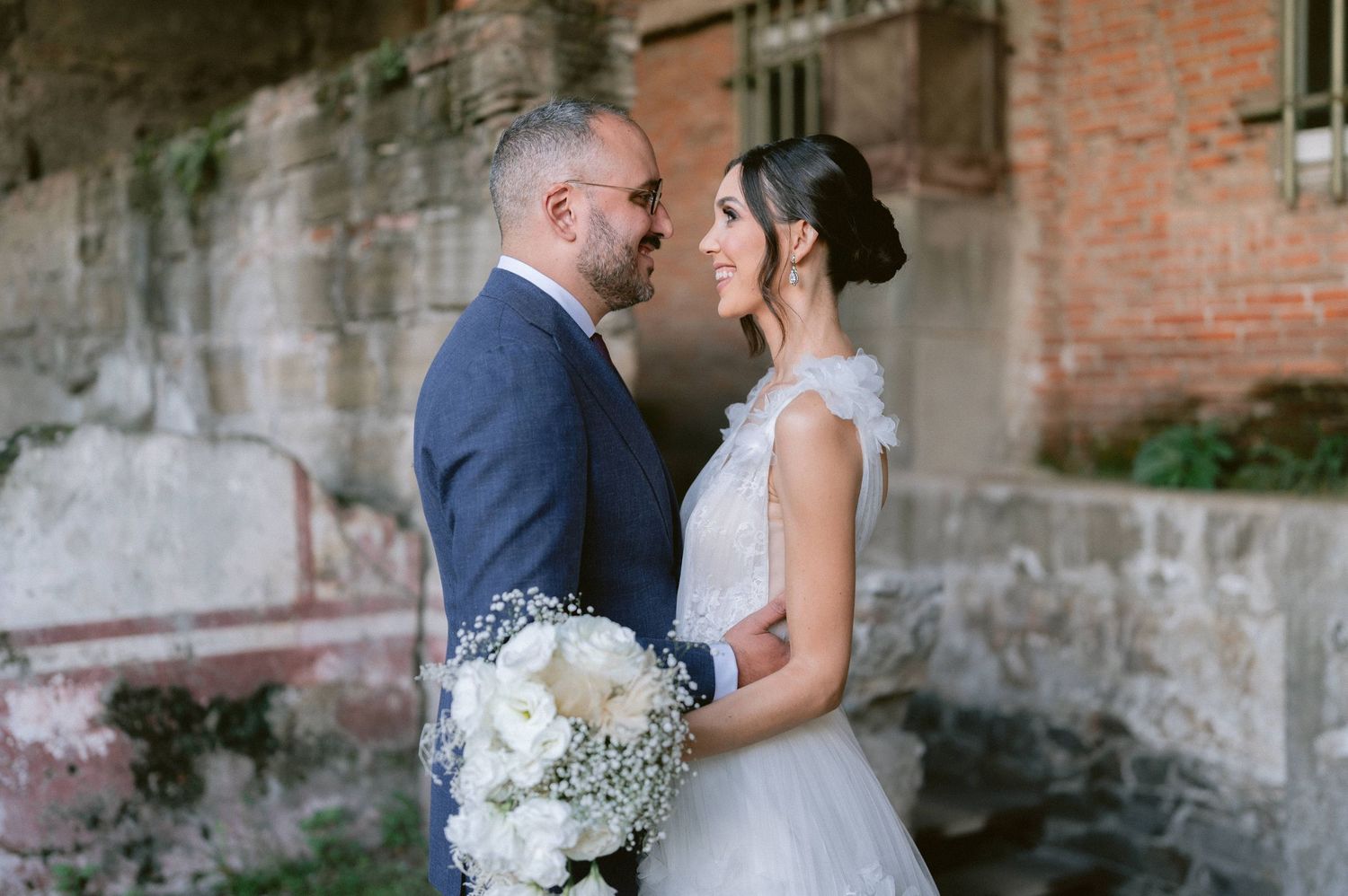 Bride and groom share an intimate moment, facing each other near rustic brick wall with white floral bouquet.