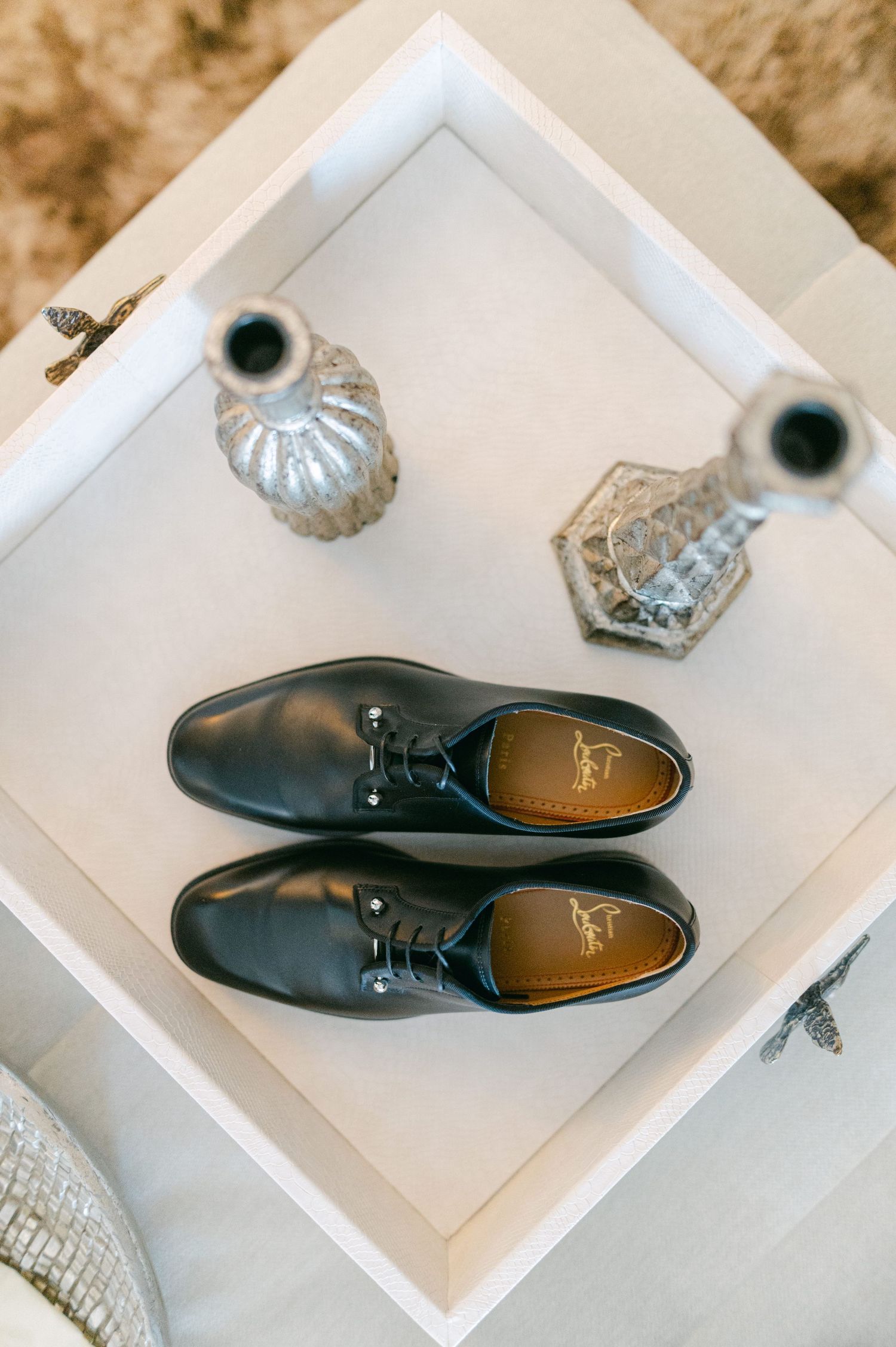 Christian Louboutin black leather Oxford dress shoes displayed on white tray with crystal vases.