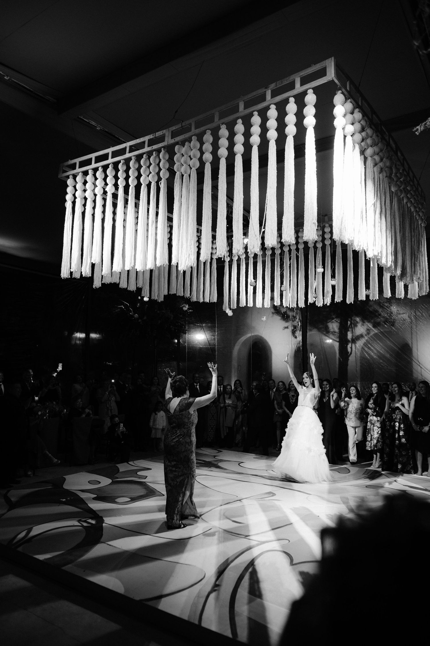 Black and white photo of bride dancing under macrame chandelier while guest faces her on dance floor.