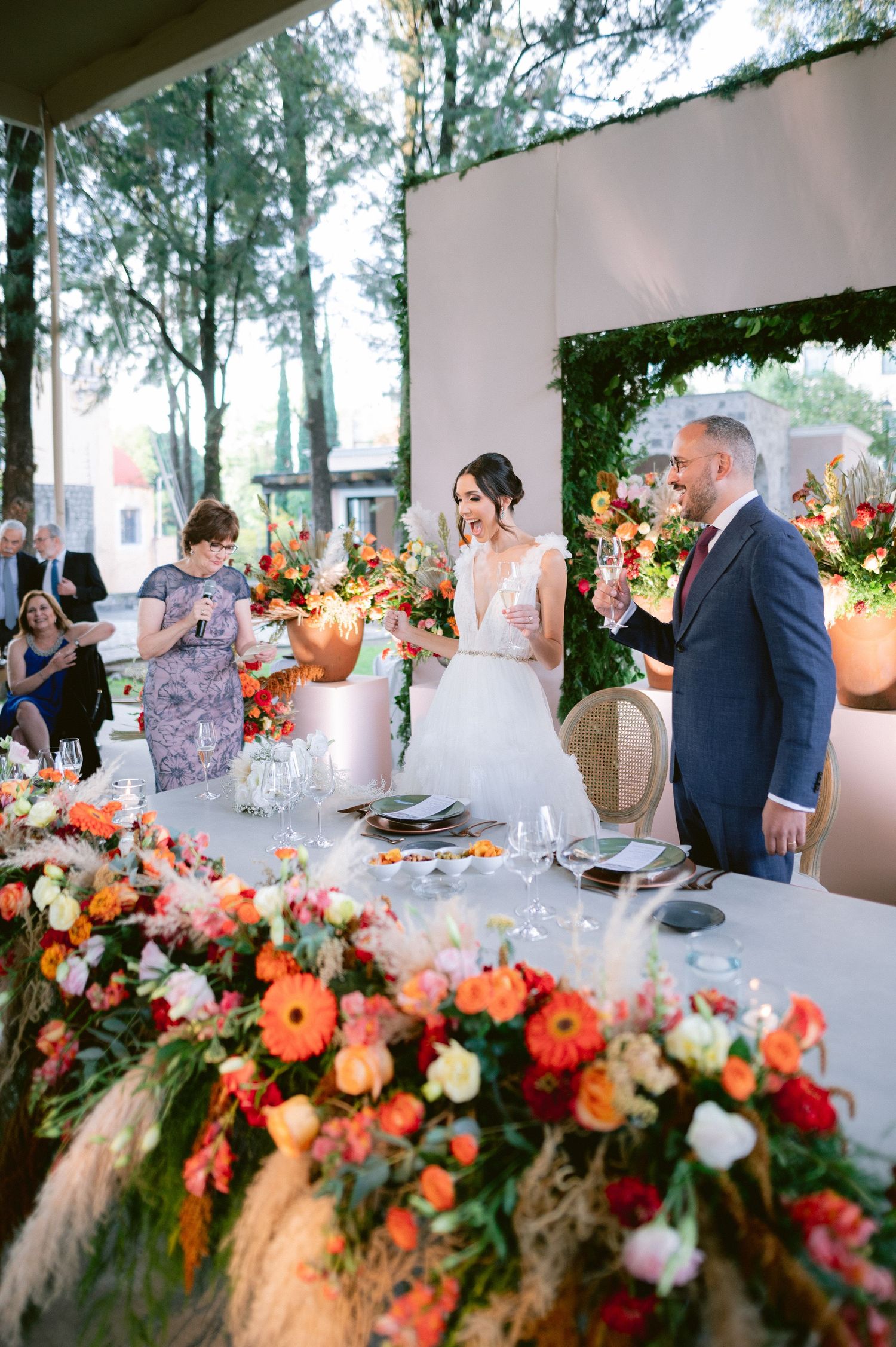 Bride and groom smile during a toast at a vibrant outdoor reception table adorned with colorful floral arrangements.