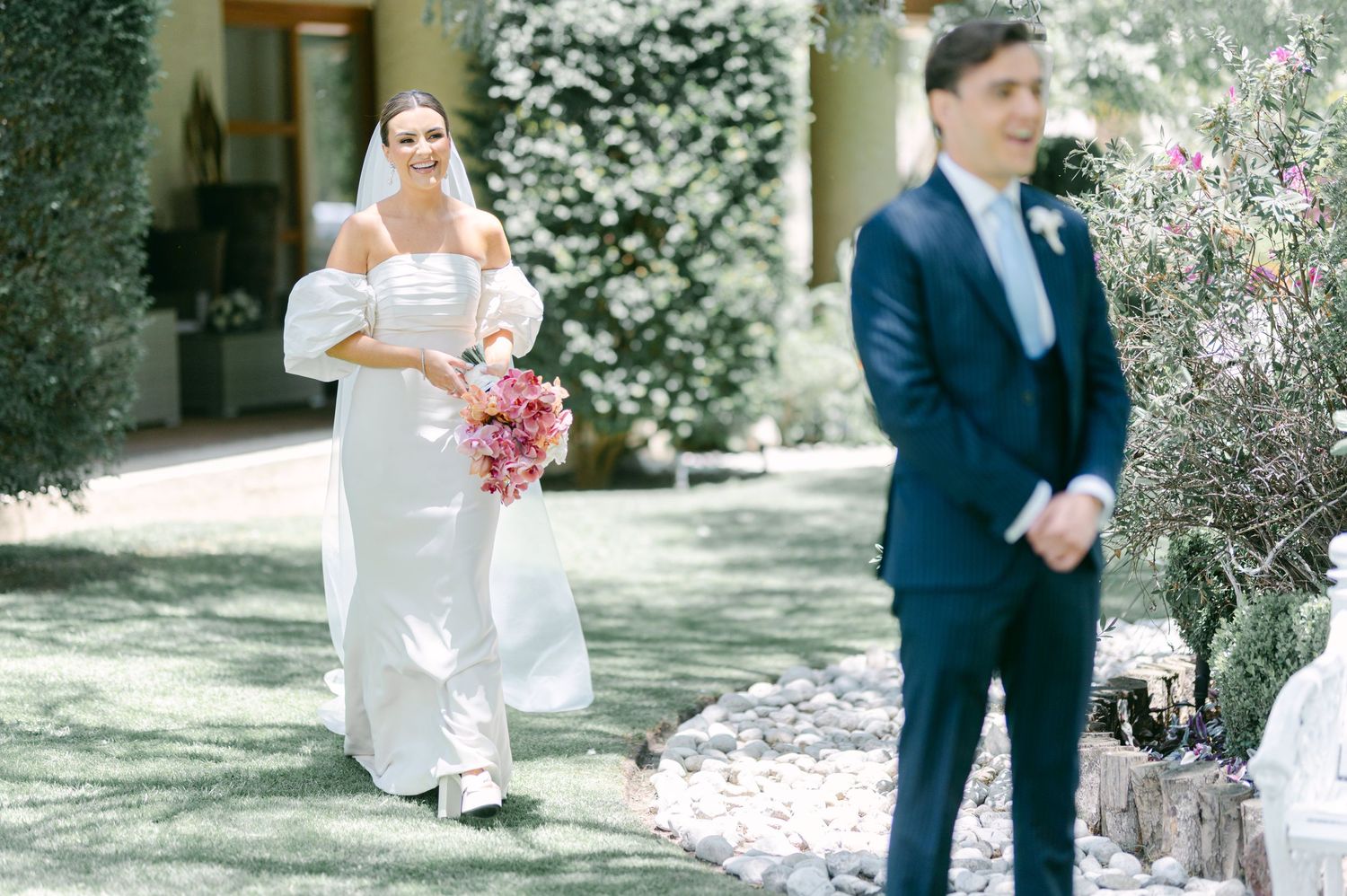 Bride in white off-shoulder gown carrying pink bouquet approaching groom outdoors.