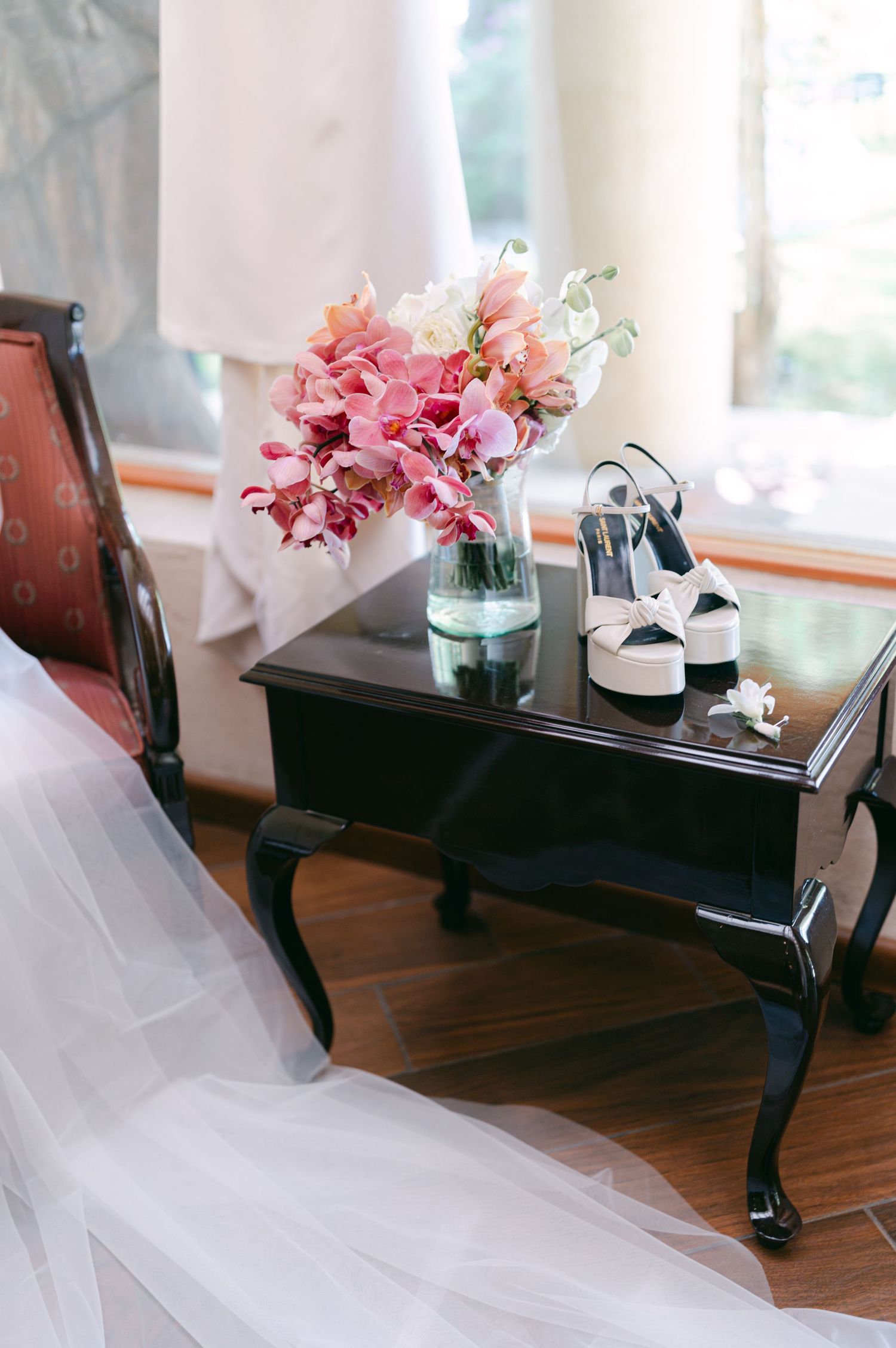 Pink orchid bridal bouquet and white heeled shoes on a black side table beside a wedding dress.