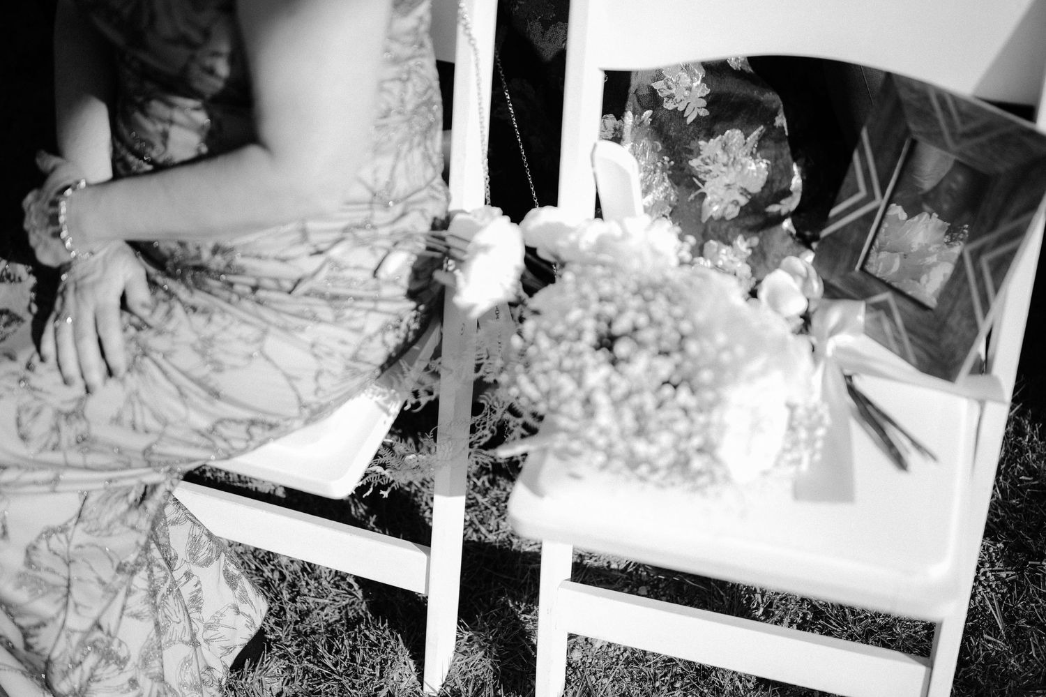 Black and white close-up of wedding guest seated in white chair holding floral bouquet at ceremony.