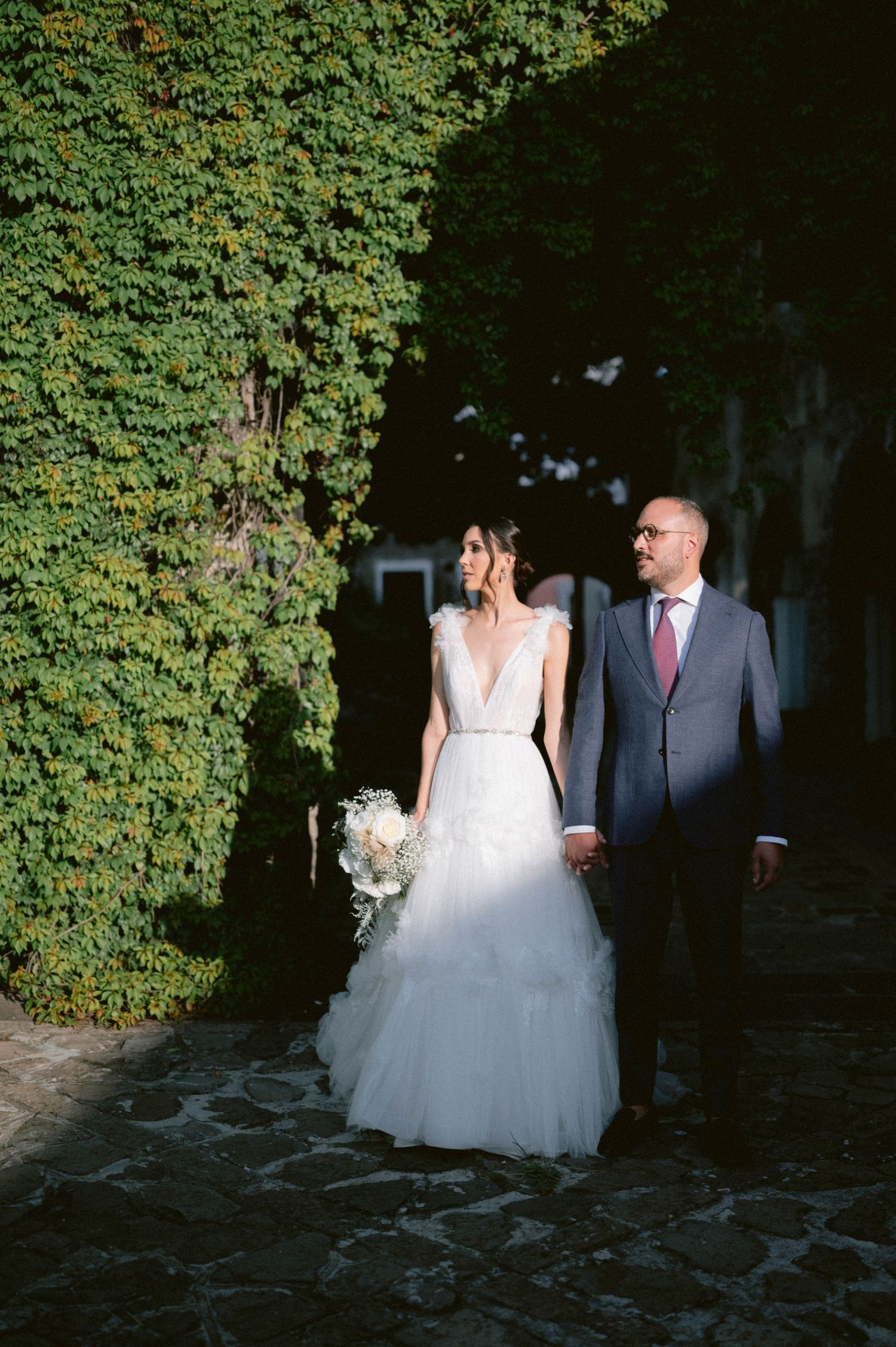 Bride holding bouquet walks with groom in blue suit along ivy-covered stone pathway.