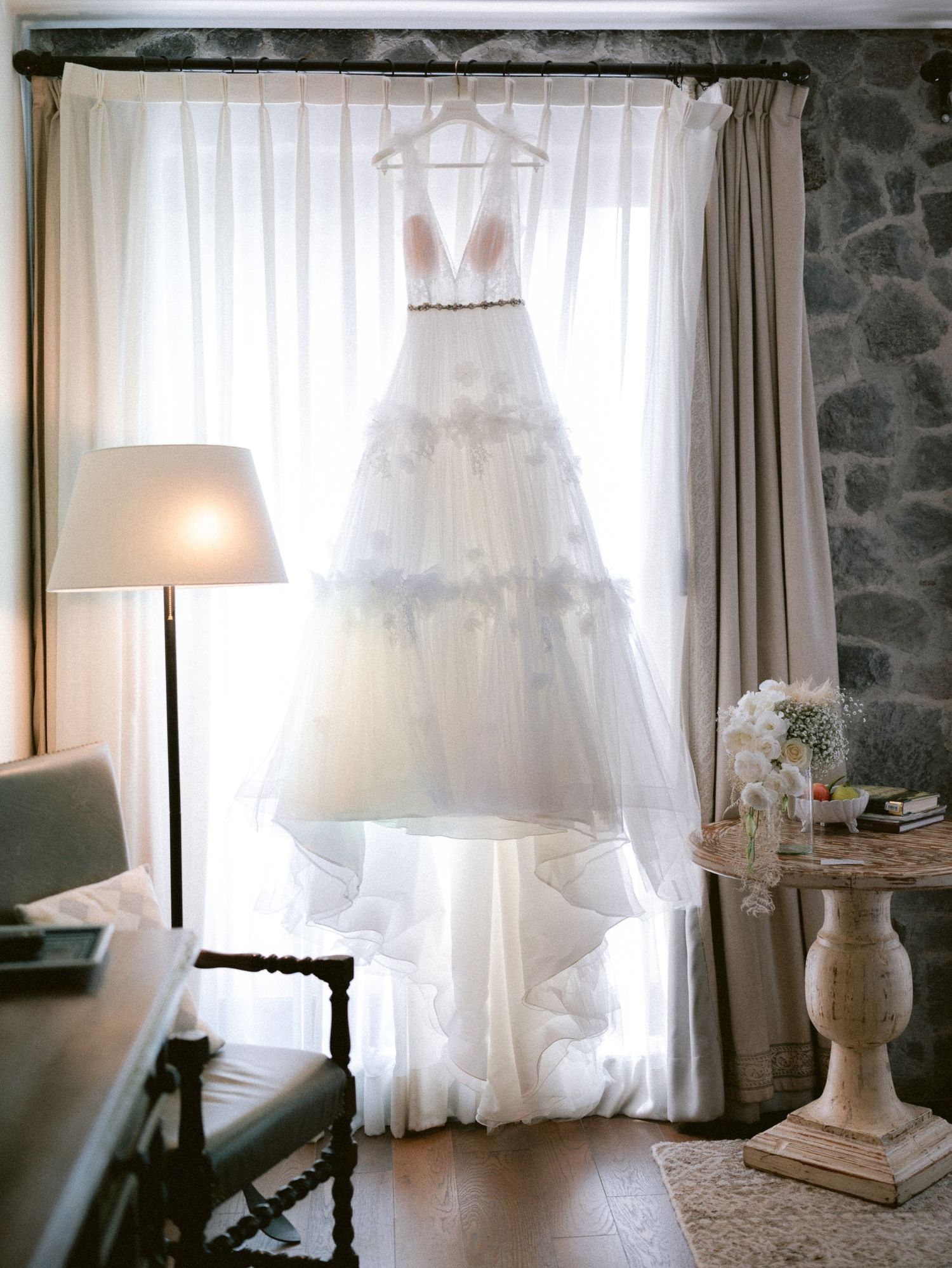 Elegant white tiered wedding gown hanging in a window with soft natural light and stone wall backdrop.