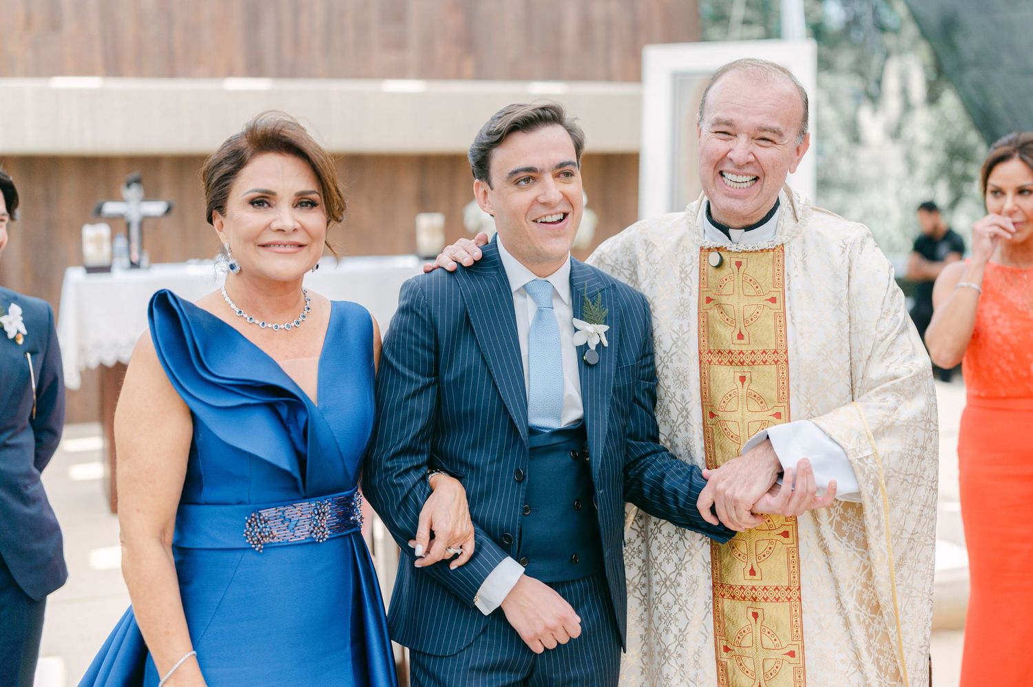Groom poses with woman in blue dress and priest after wedding ceremony outdoors.