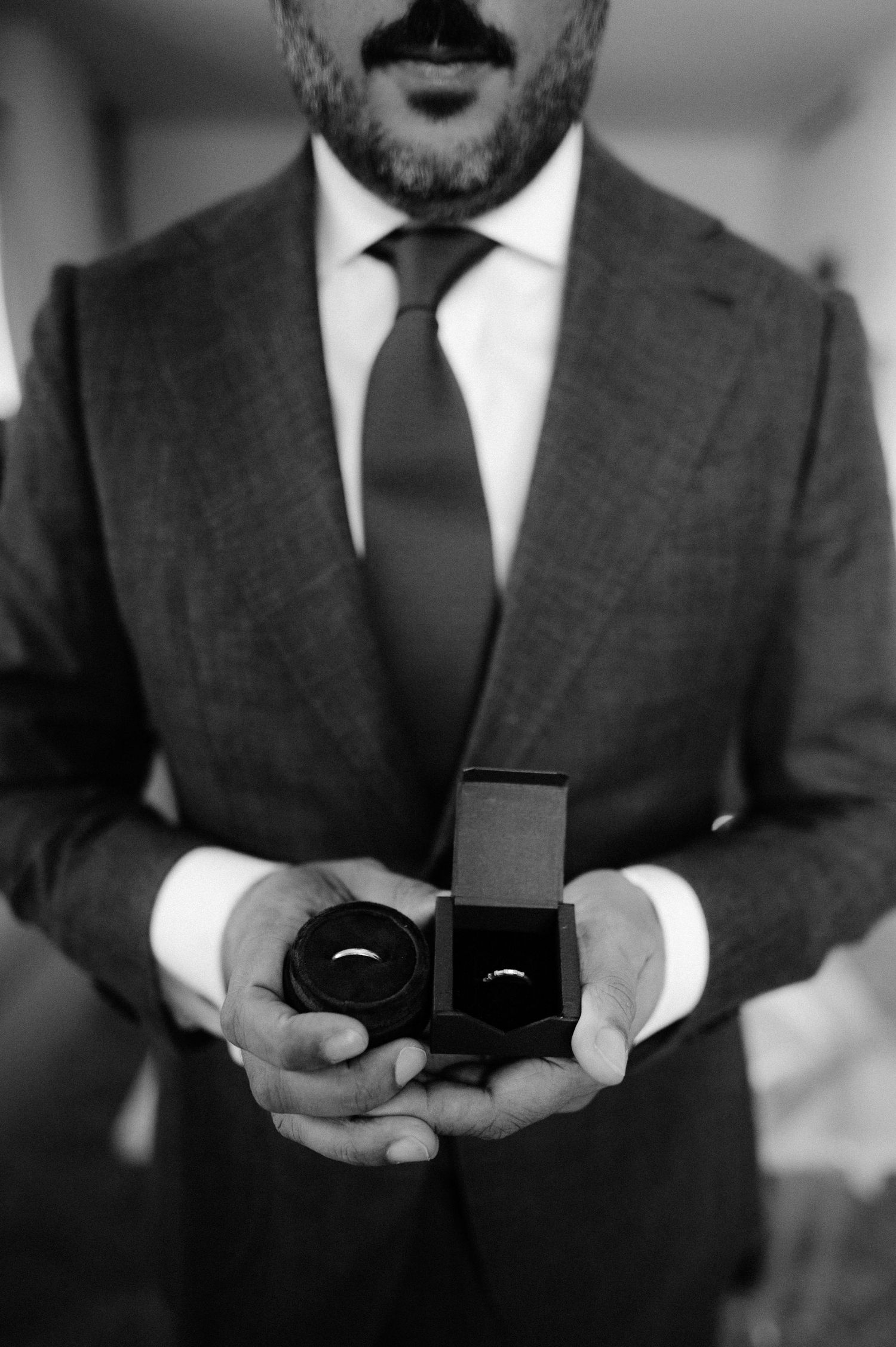 Black and white photo of groom in suit holding two wedding rings in velvet boxes.