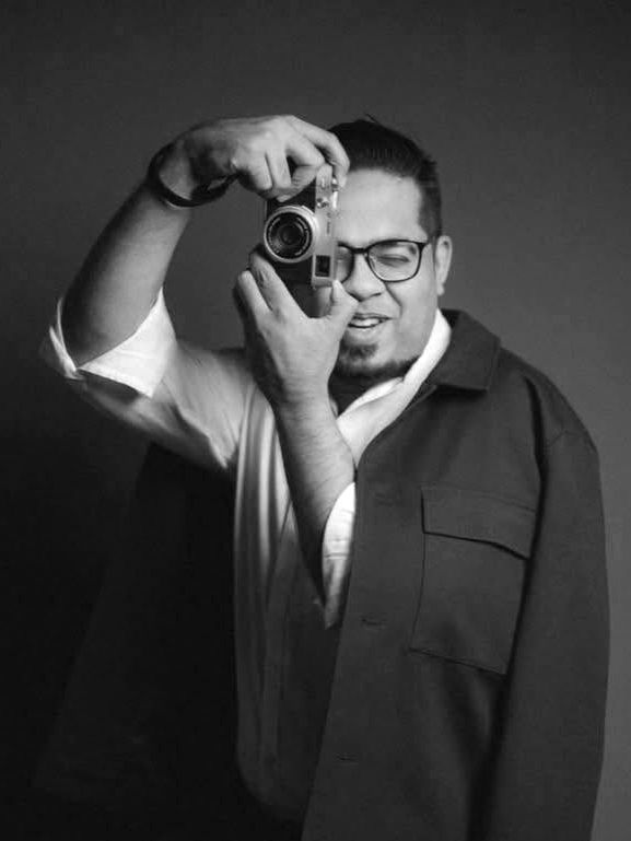 Man in glasses holding a vintage camera up to his eye in a black and white portrait photo.